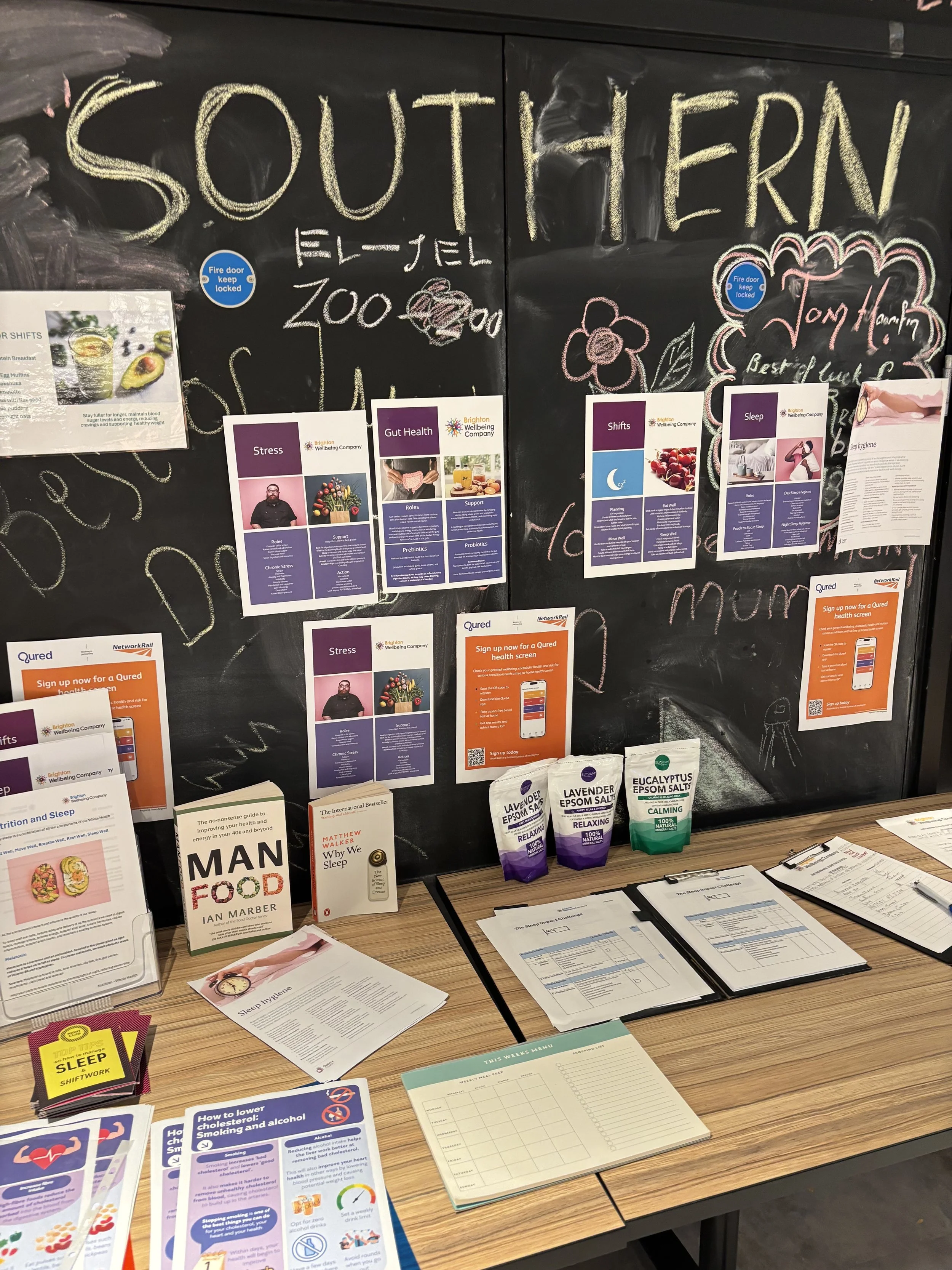 Blackboard with colorful chalk writing, printed health and wellness flyers, booklets, pamphlets, paper notepads, and supplements on a wooden table in front.