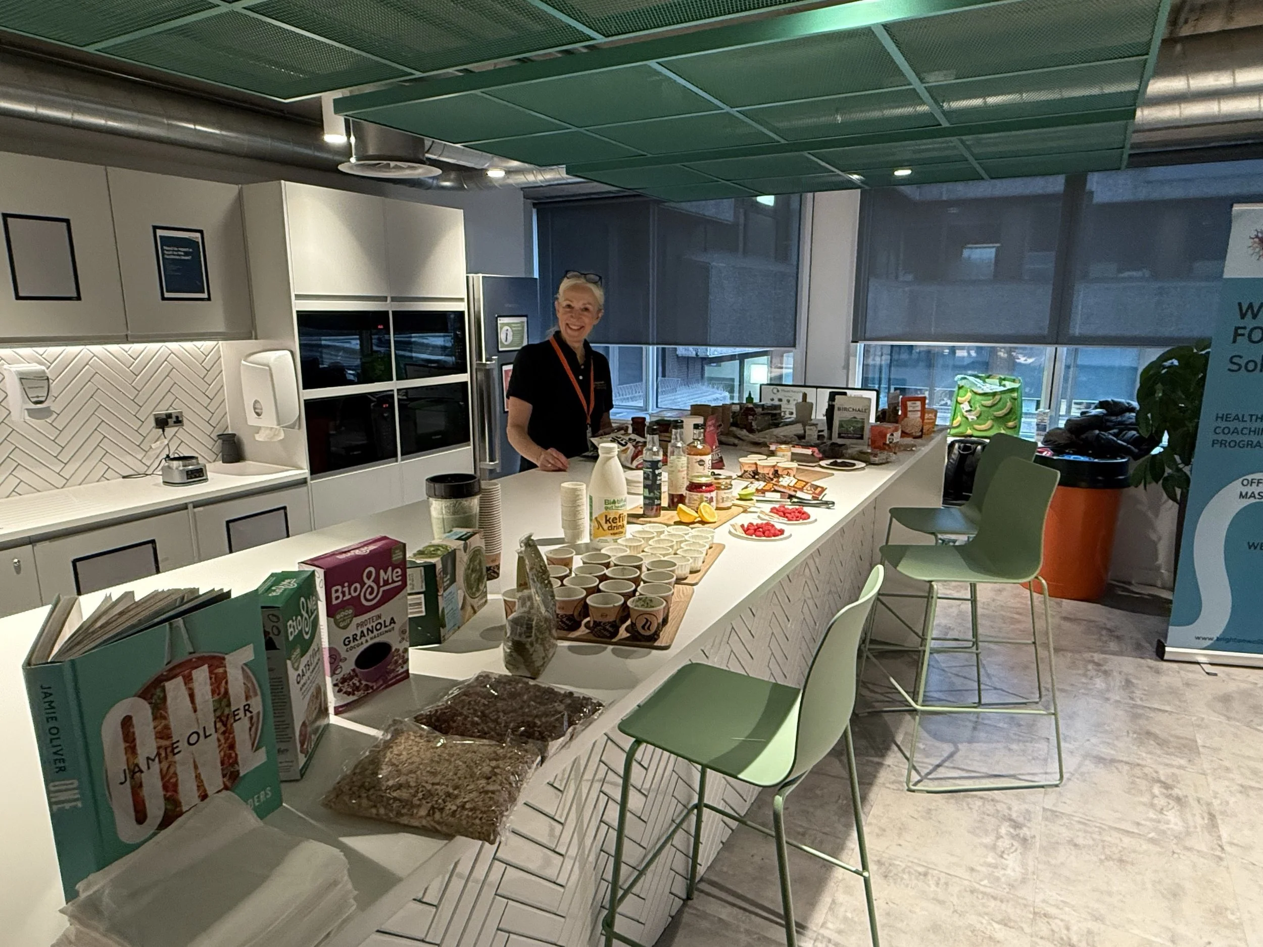 A woman standing behind a white breakfast bar with various food items, drinks, and breakfast products in a modern kitchen or cafeteria.