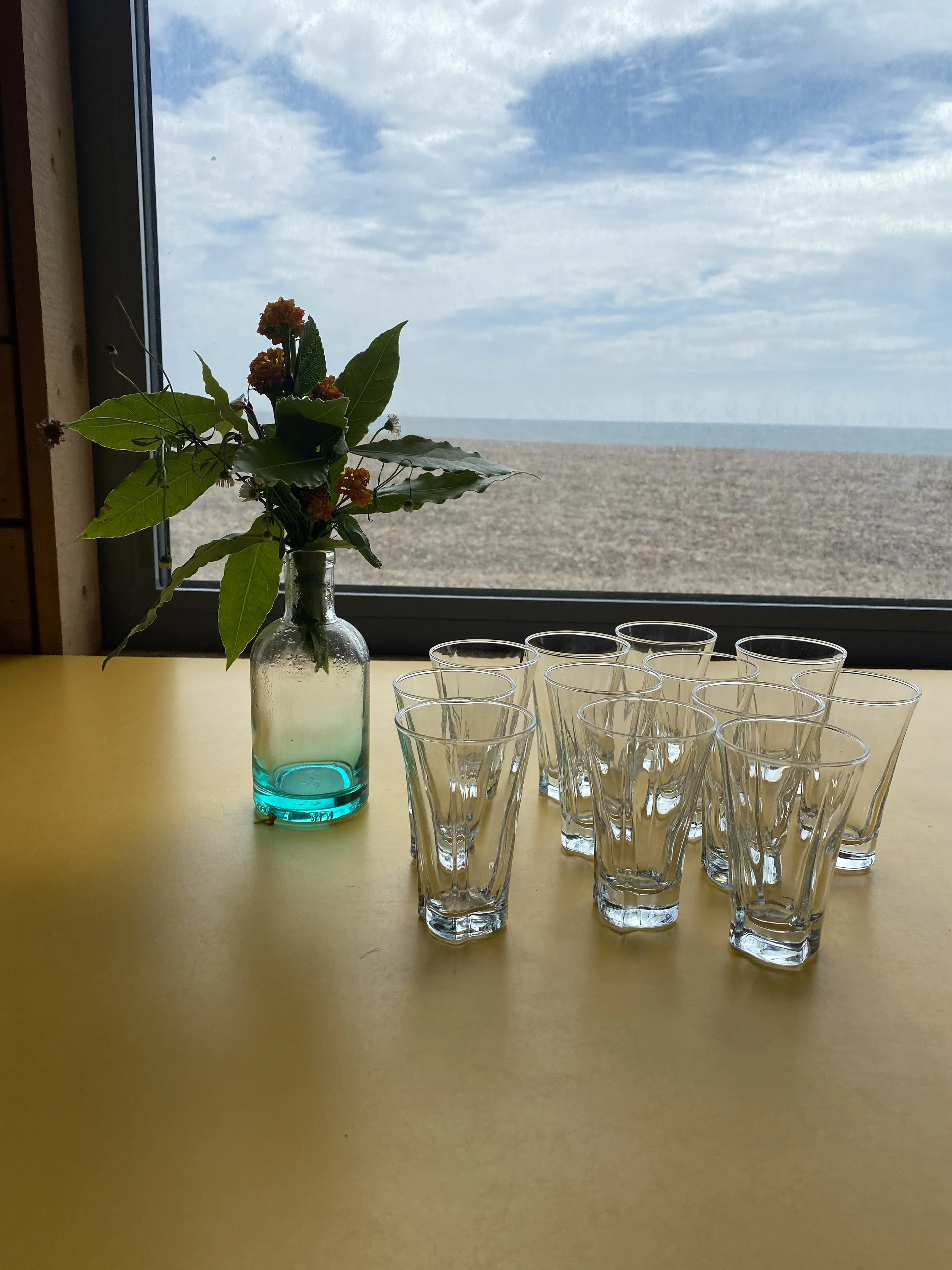 A vase with flowers and multiple glasses on a yellow table with a view of the beach and sky outside the window.