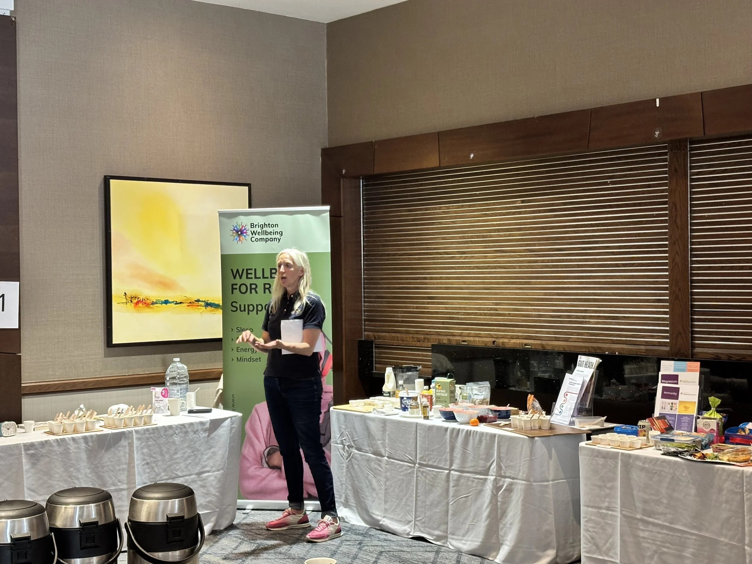 A woman standing in front of a table with food and promotional materials at an event, with a Brighton Wellbeing Company banner behind her.