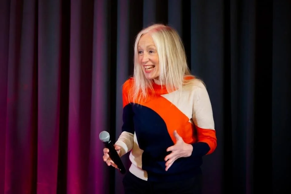 A woman with long blonde hair holding a microphone, smiling, standing in front of dark curtains.