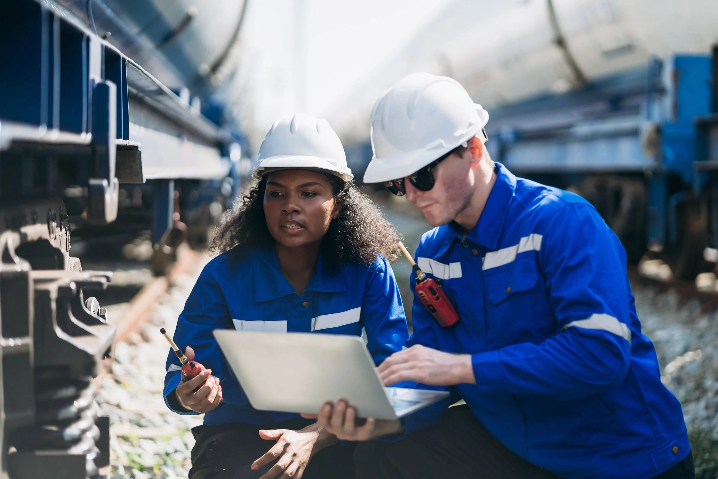 Two electrical workers, a woman and a man, wearing blue uniforms, white hard hats, and safety glasses, examining equipment on a train track with a laptop and walkie-talkie.