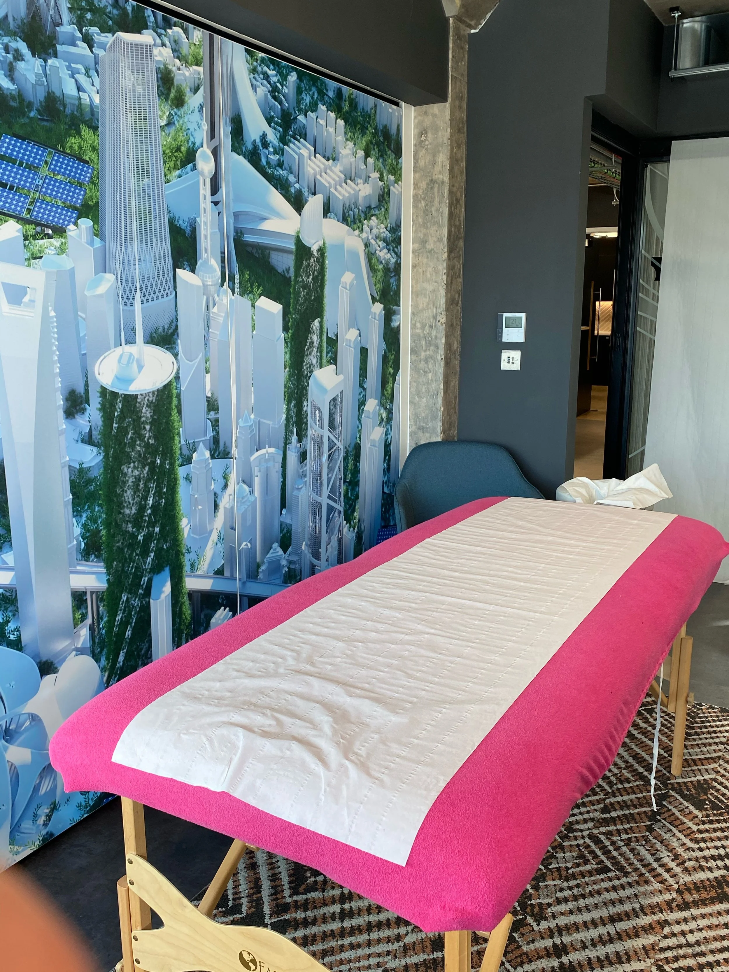 Room with a massage table covered with a pink blanket and a white disposable sheet, next to a black chair and a colorful rug, with a large wall mural of a futuristic cityscape featuring tall, white skyscrapers and flying vehicles.