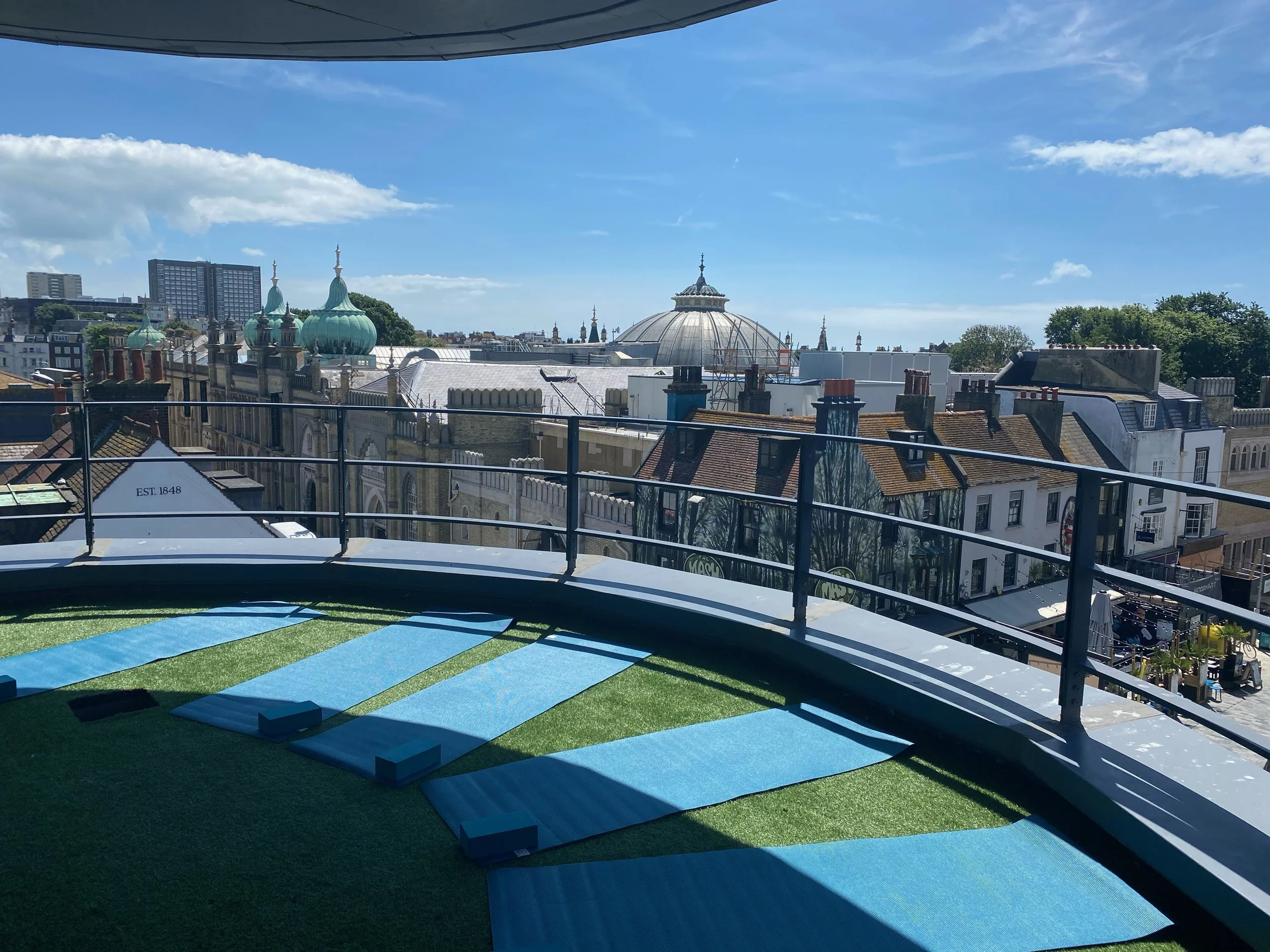 Rooftop with four blue yoga mats on artificial grass, overlooking historic and modern buildings with domed and spired rooftops under a clear blue sky.