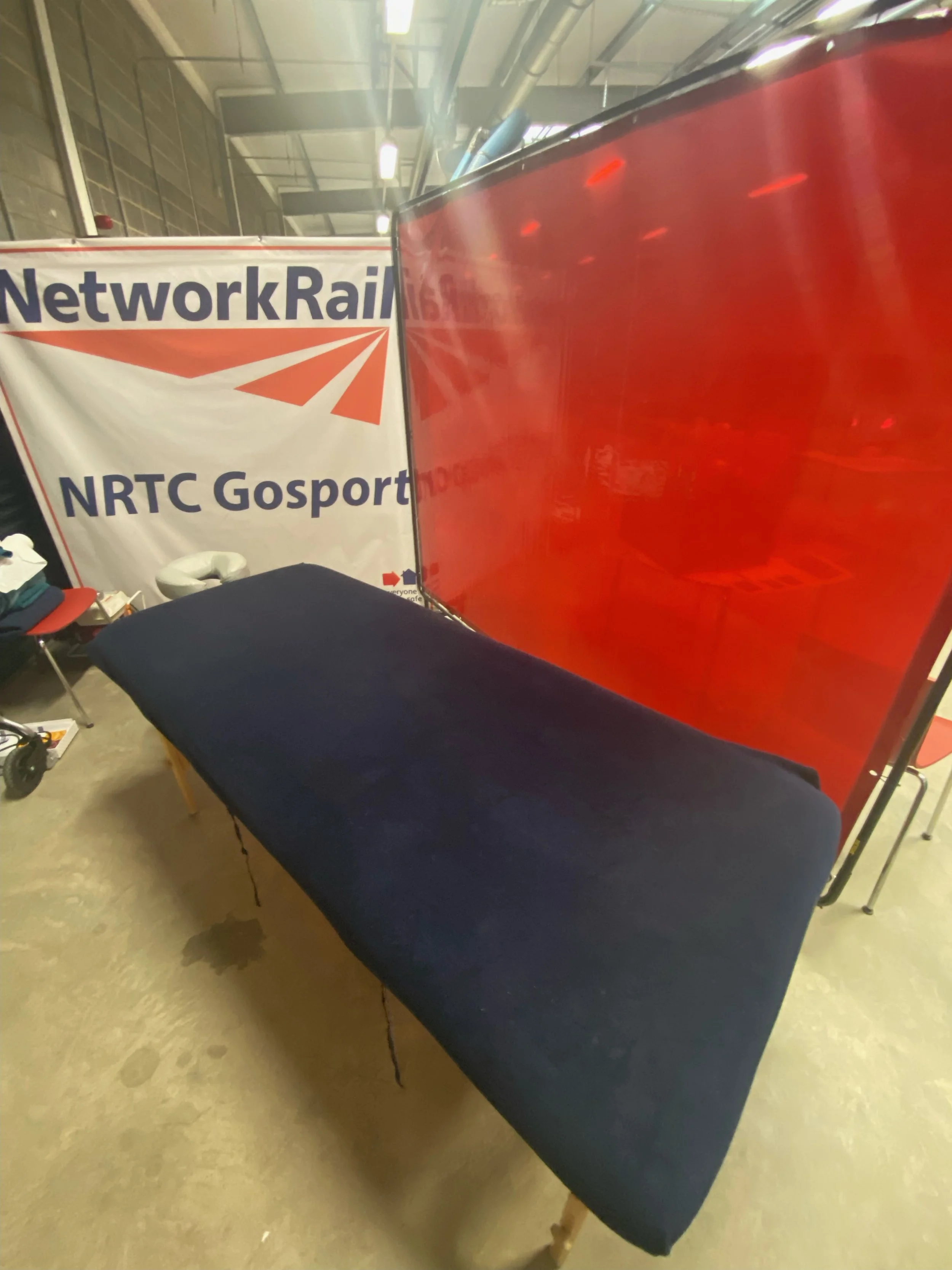 An indoor scene with a black massage table, a red partition, and a banner that reads "Network Rail" and "NRTC Gosport."