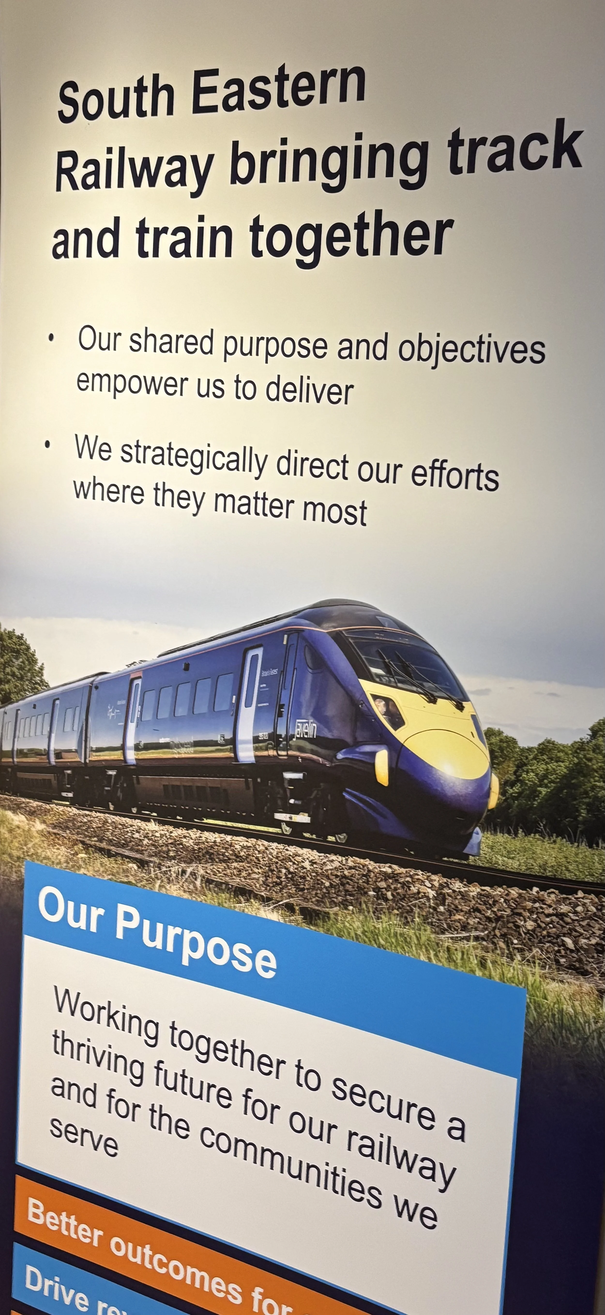 A poster featuring a blue and yellow modern train on the tracks. The background shows a cloudy sky and green trees. The poster contains text about South Eastern Railway's purpose and objectives, emphasizing teamwork and community service.