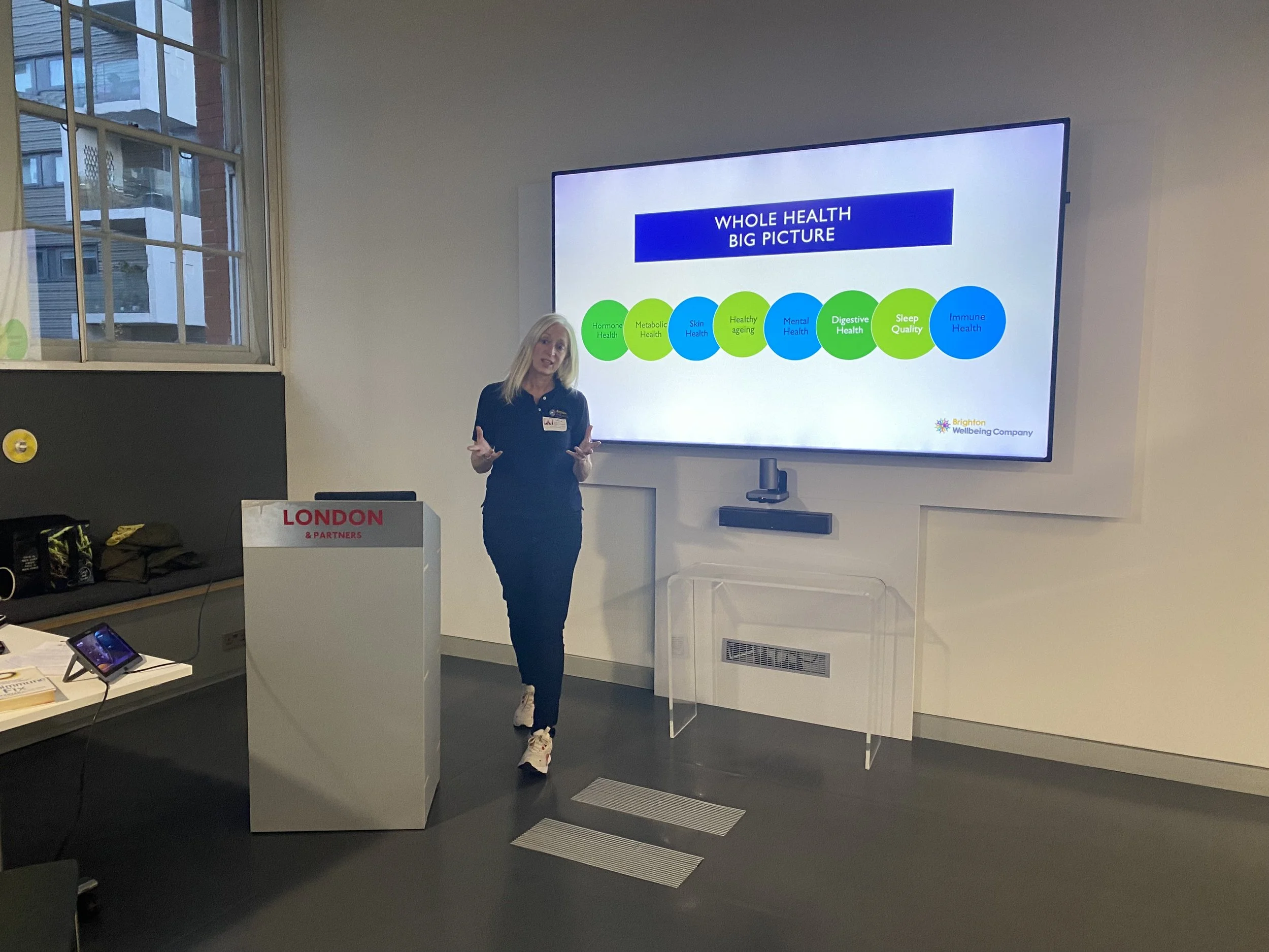 A woman giving a presentation in a conference room with a large screen behind her displaying a slide titled 'Whole Health Big Picture'. The slide features a row of colored circles representing different health aspects, including Hormone Health, Metab