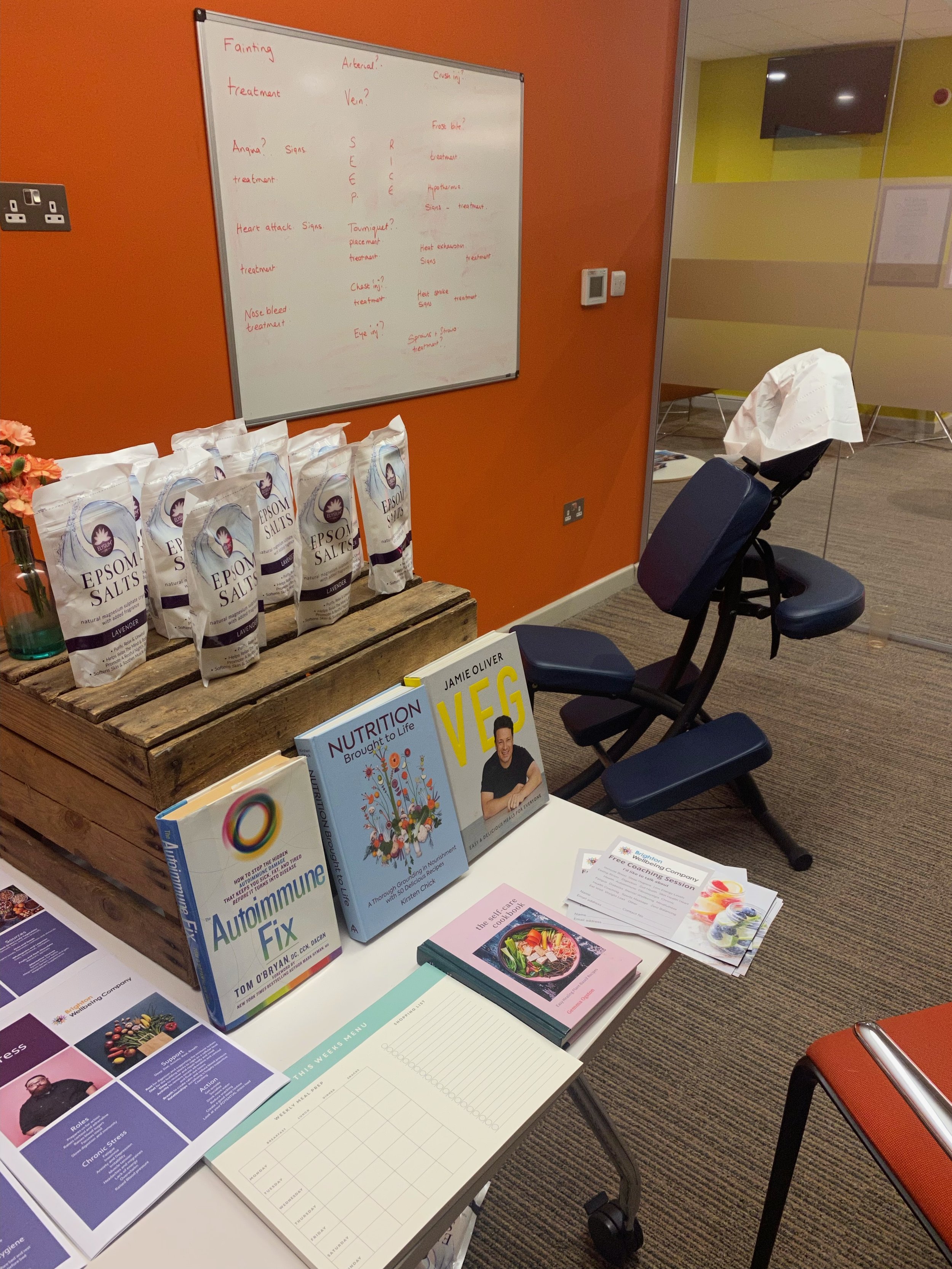 Display table with health and nutrition books, packets of Epsom salts, posters, and informational materials, set up in a conference room with orange and glass walls, a whiteboard with handwritten notes, and a massage chair.