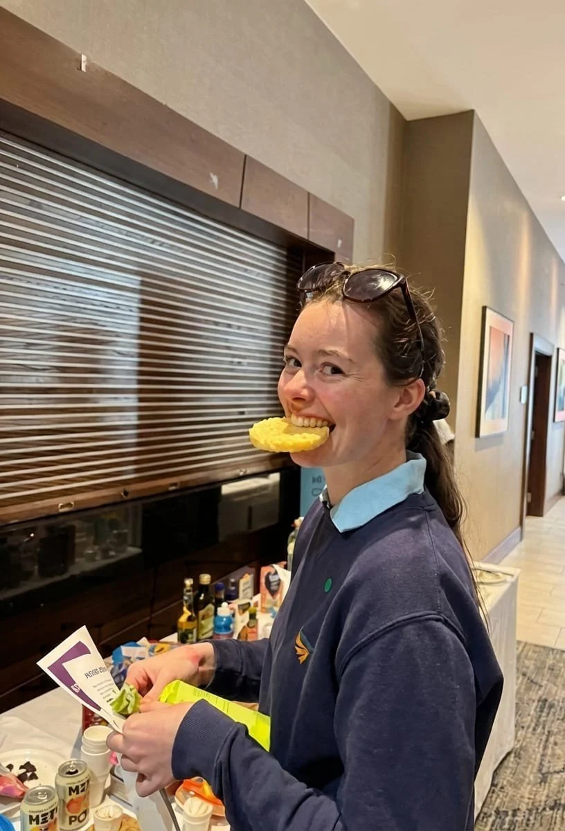 Woman with glasses on her head biting into a pineapple ring at a snack table in an indoor setting.