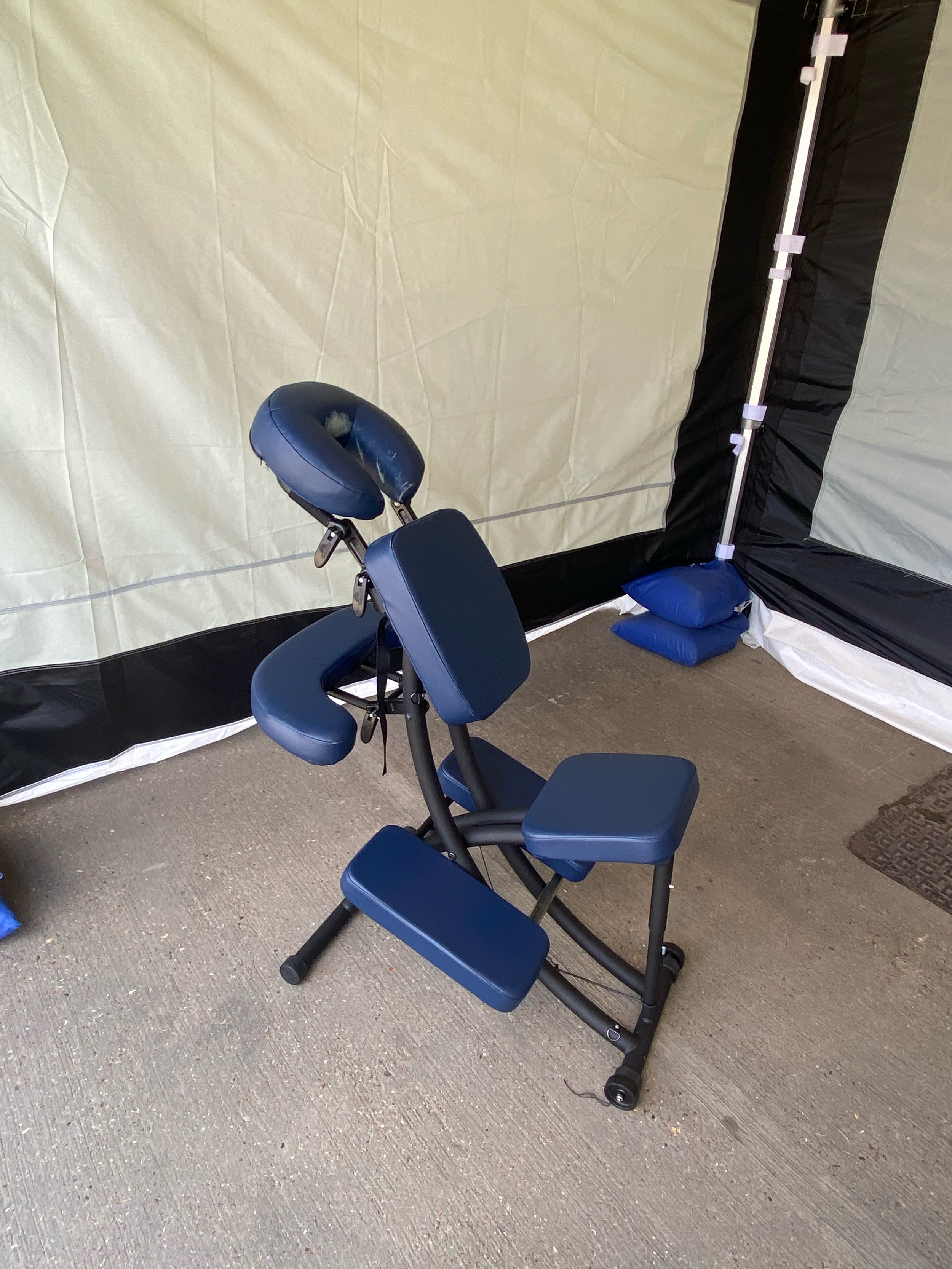 Adjustable massage chair with blue upholstery inside a medical or therapy tent with beige and black walls, placed on a concrete floor.