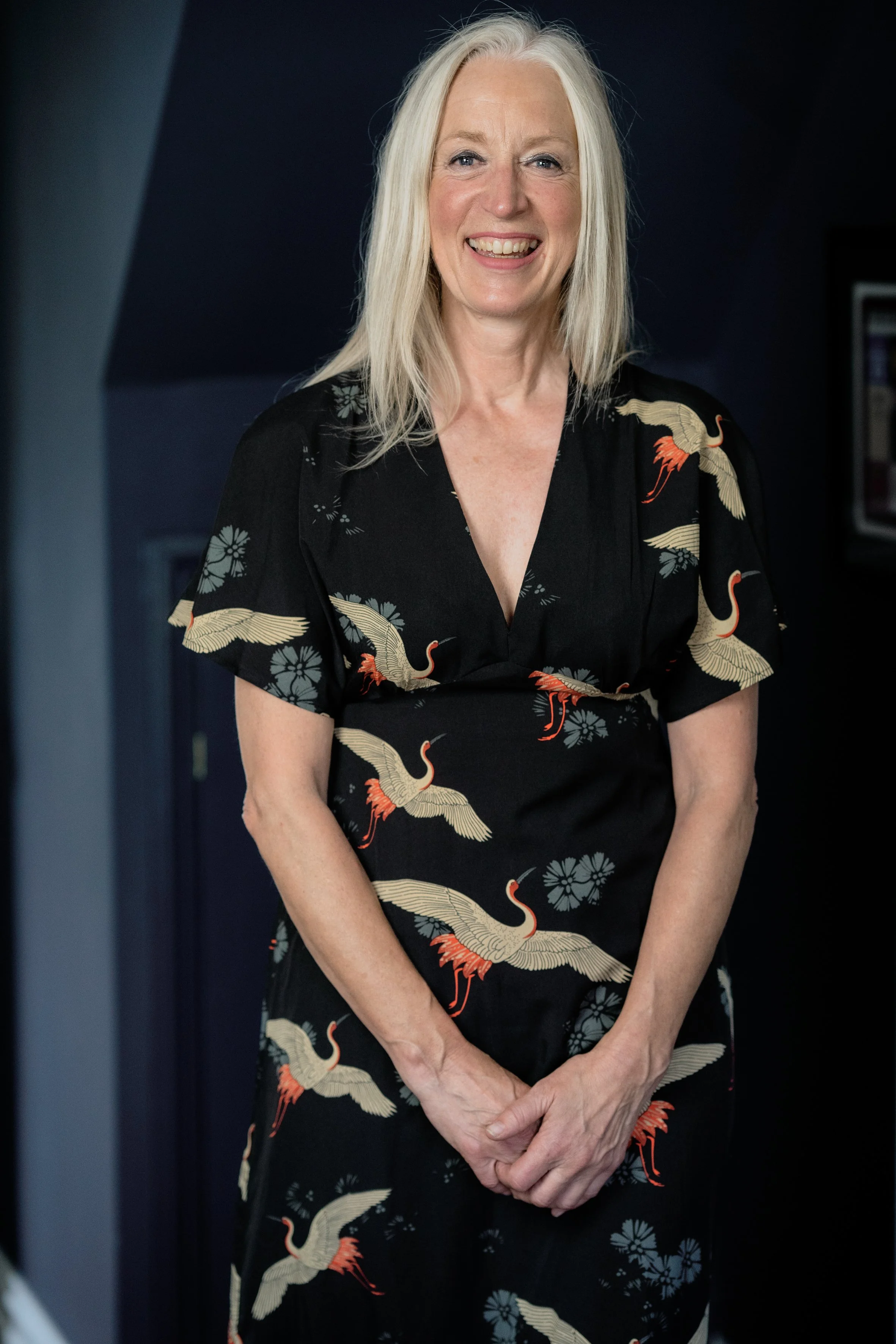 A smiling woman with long, gray hair standing indoors in front of a dark background, wearing a black dress with crane and floral patterns.
