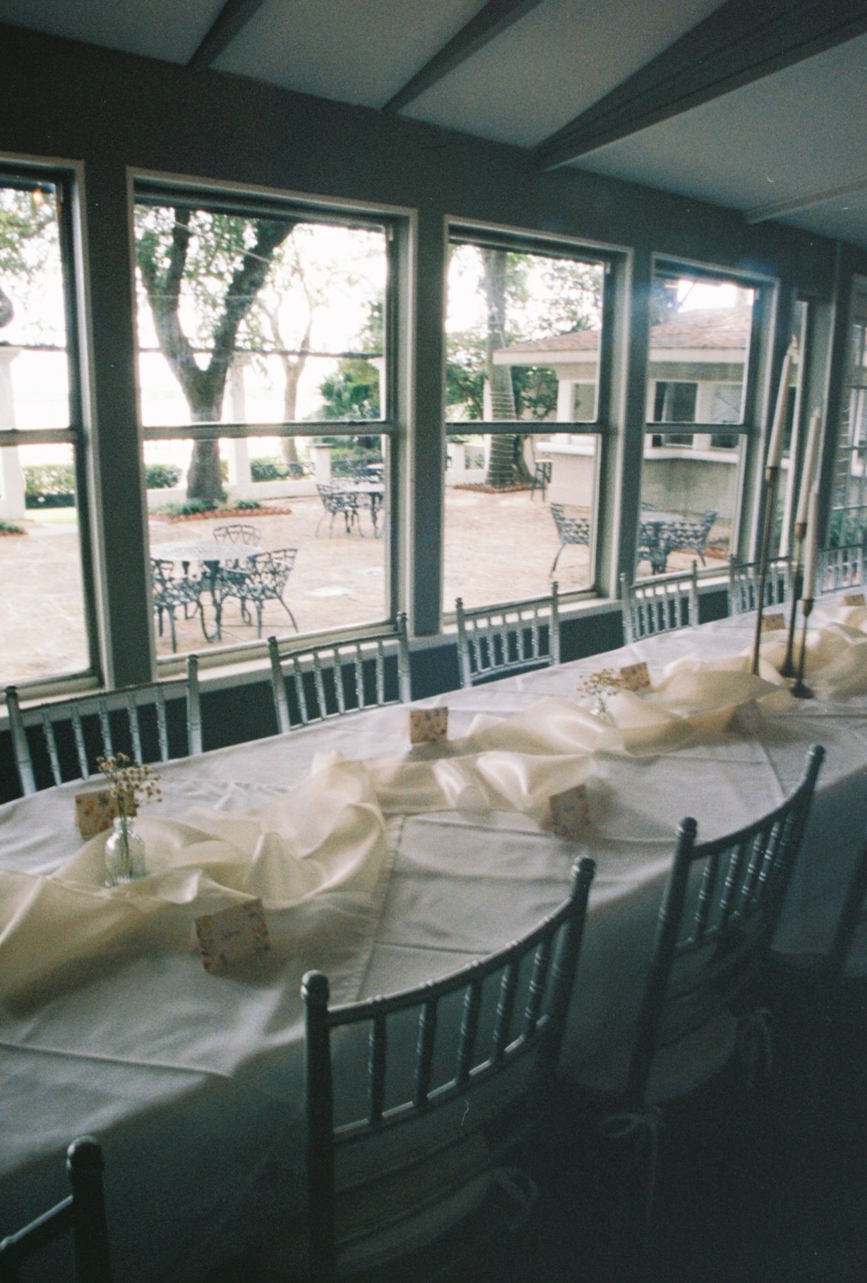 Indoor dining area set up for a gathering with a long table covered with a white tablecloth, decorated with folded napkins and small floral centerpieces. Large windows reveal an outdoor patio with several tables and chairs, and trees in the backgroun