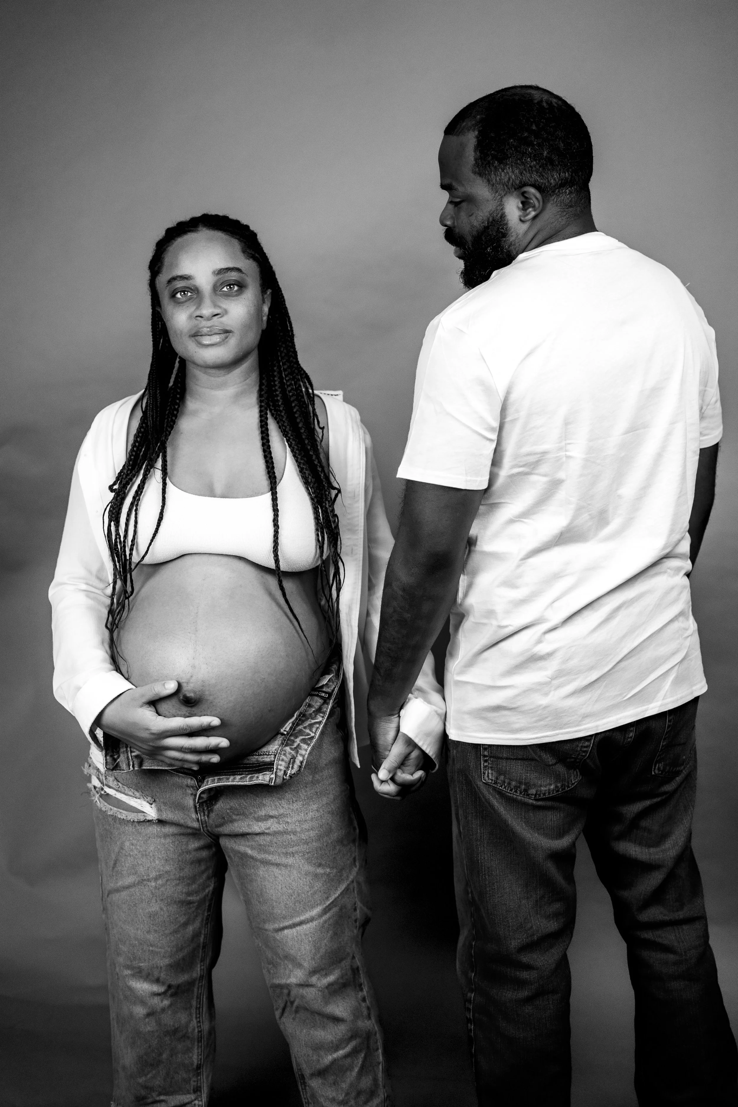 A pregnant woman with long braids, wearing a white top and ripped jeans, stands holding her belly, while a man in a white t-shirt and jeans holds her hand, both standing against a plain background.