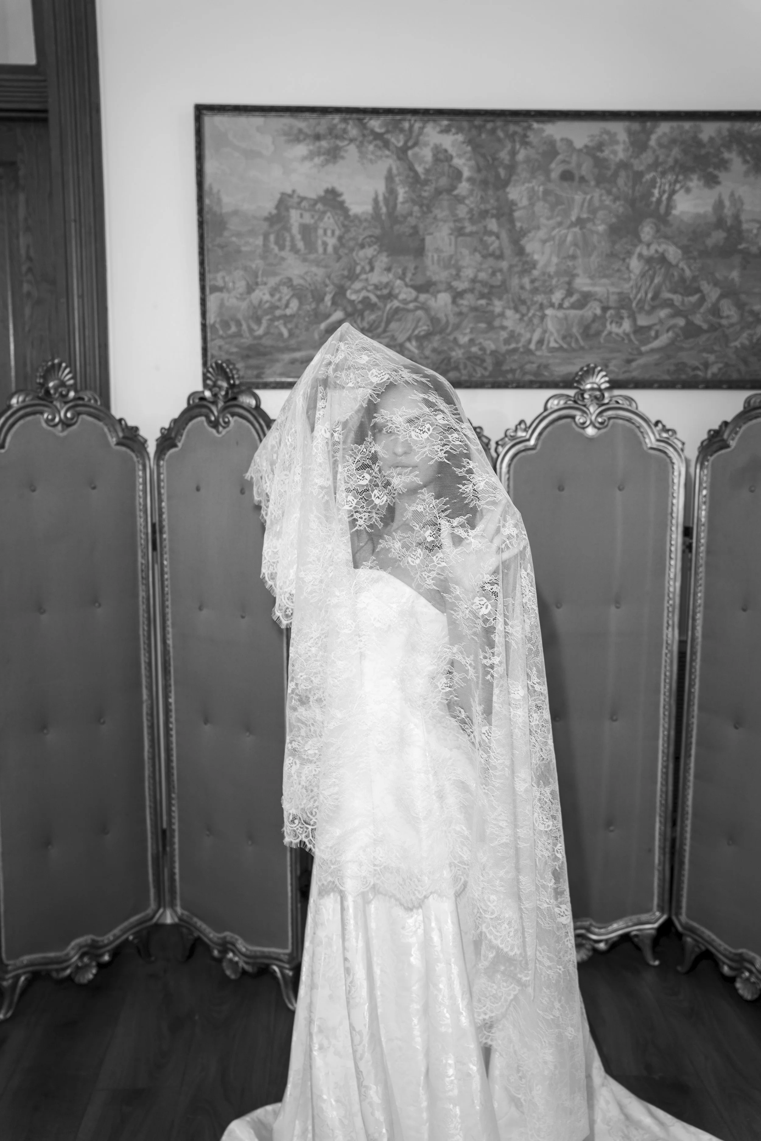 A vintage wedding dress with lace details is draped over a chair in a room with wooden floors, a large framed tapestry on the wall, and ornate room dividers.