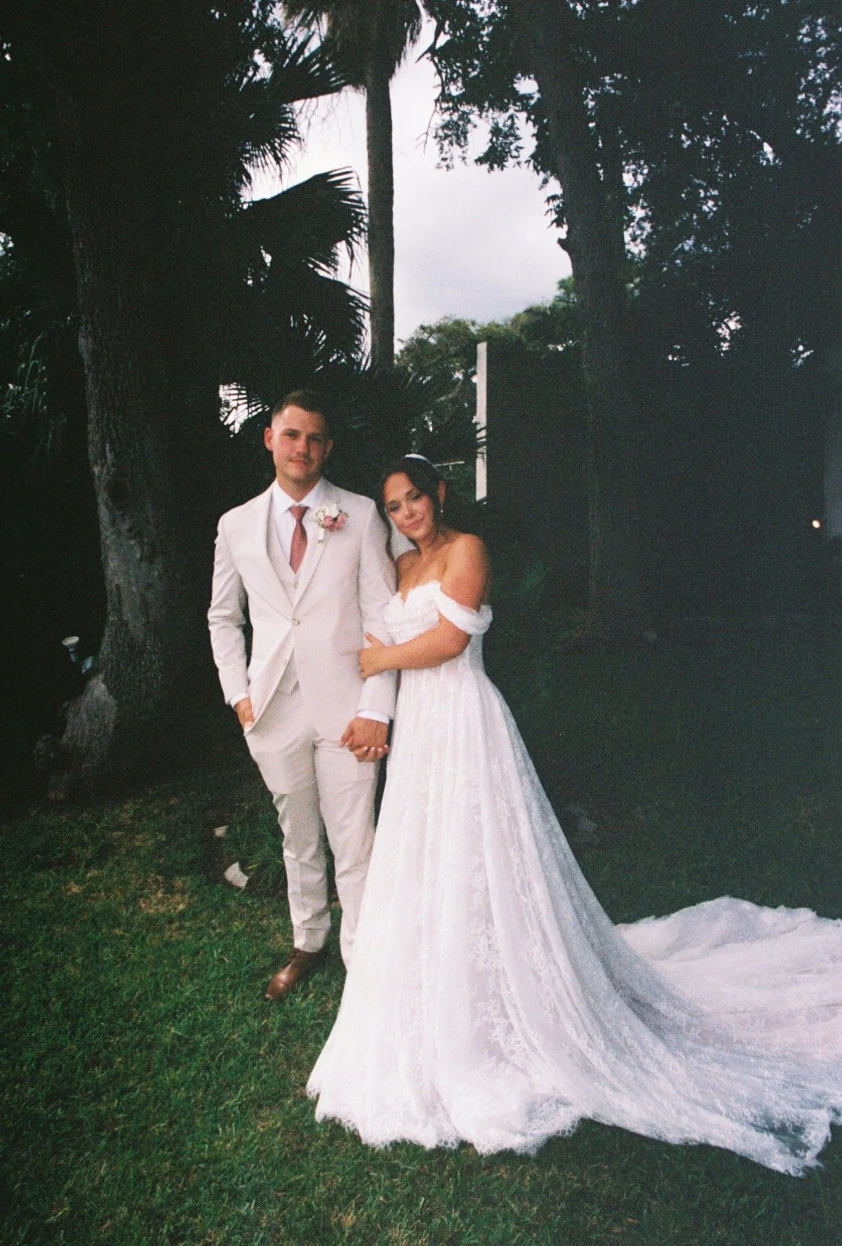 A bride and groom standing outdoors in front of large trees during their wedding, holding hands, with the bride wearing a white wedding gown and the groom in a light-colored suit.
