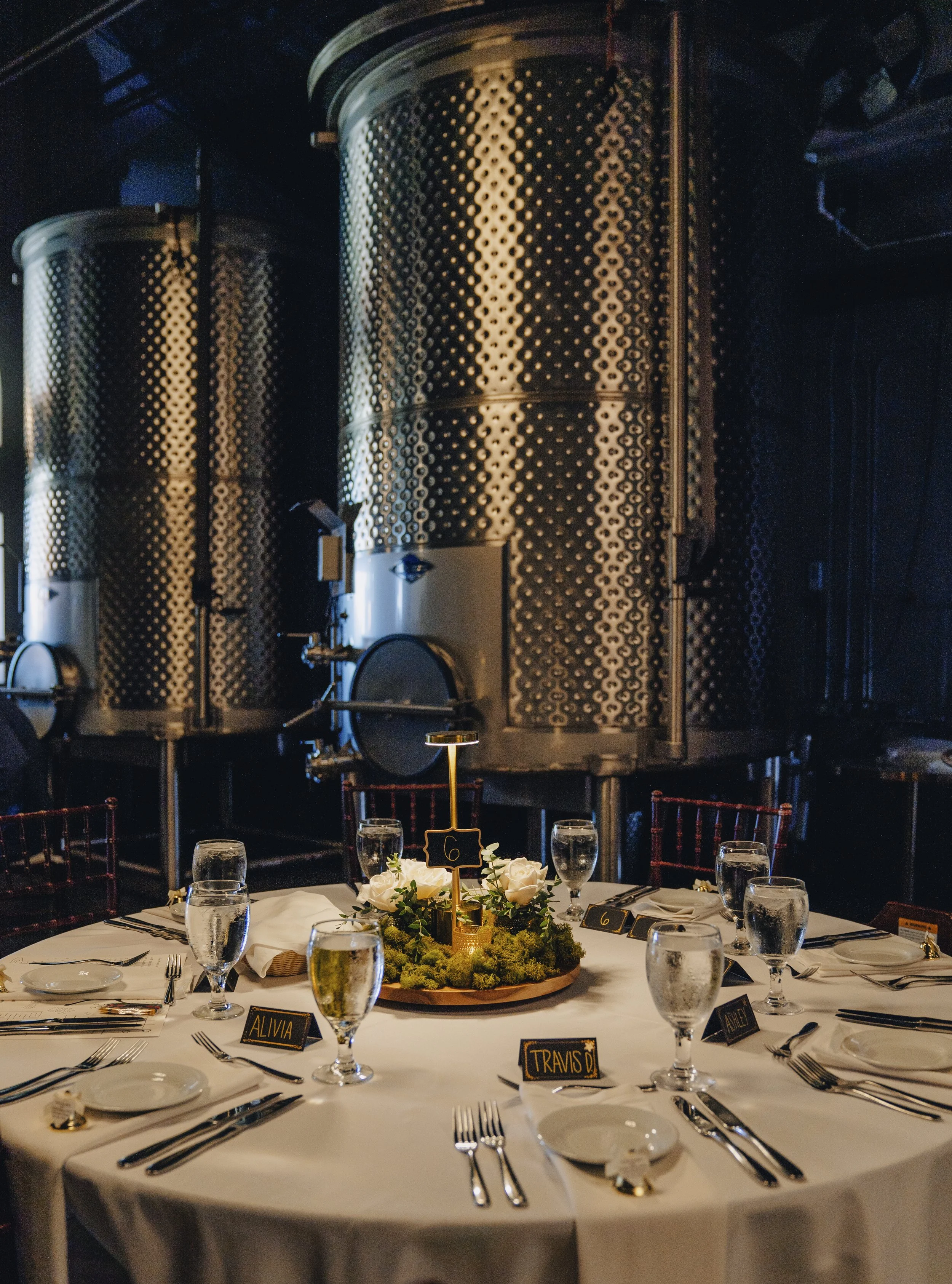Elegantly set round dinner table with a white tablecloth, glassware, plates, silverware, and small black place cards with gold text, in a dimly lit industrial-style venue. The centerpiece is a floral arrangement with white roses and greenery, and the