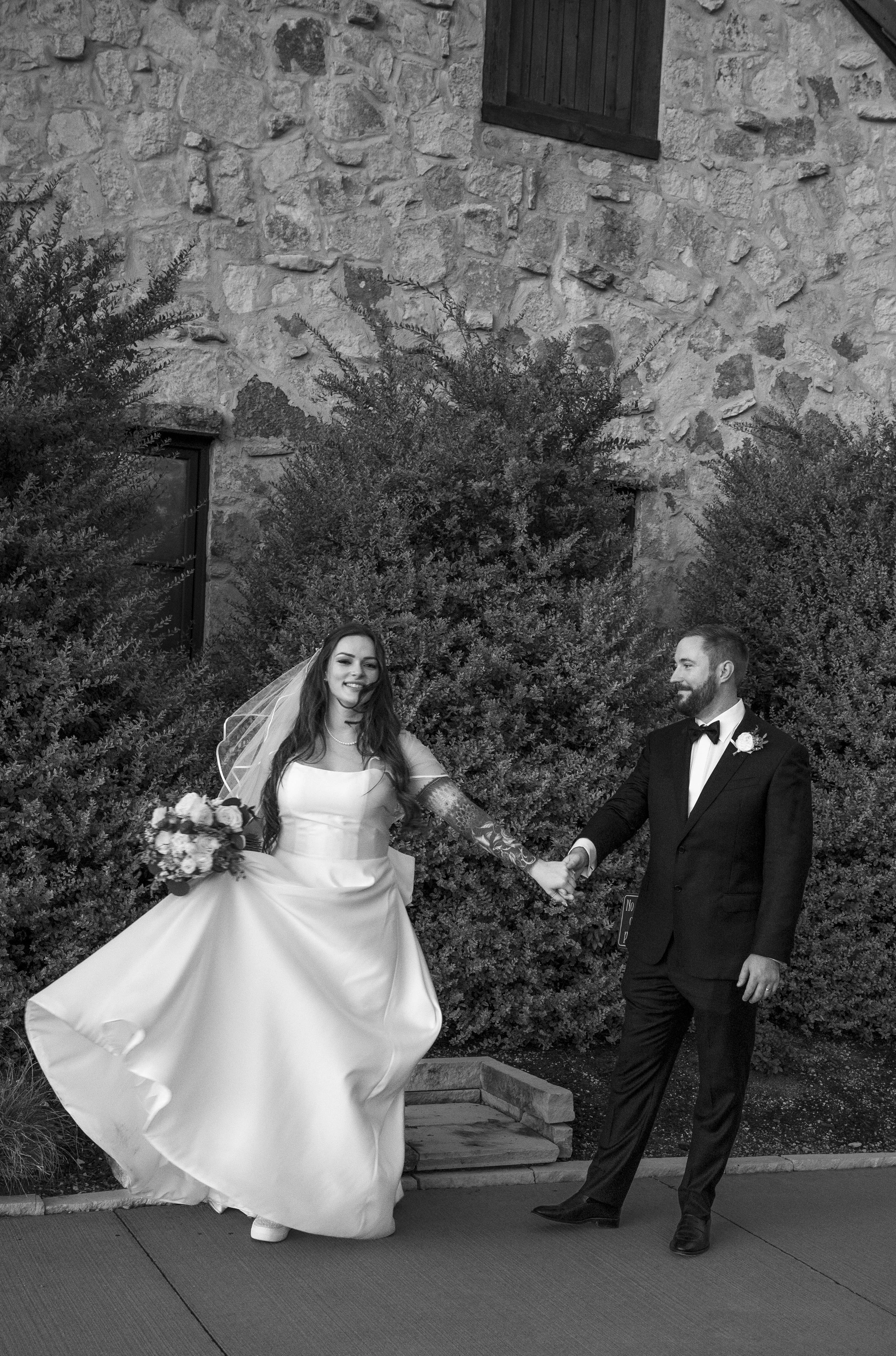 A bride and groom holding hands outdoors, with the bride in a wedding dress and veil, and the groom in a tuxedo with a boutonnière, standing in front of bushes and a stone building.