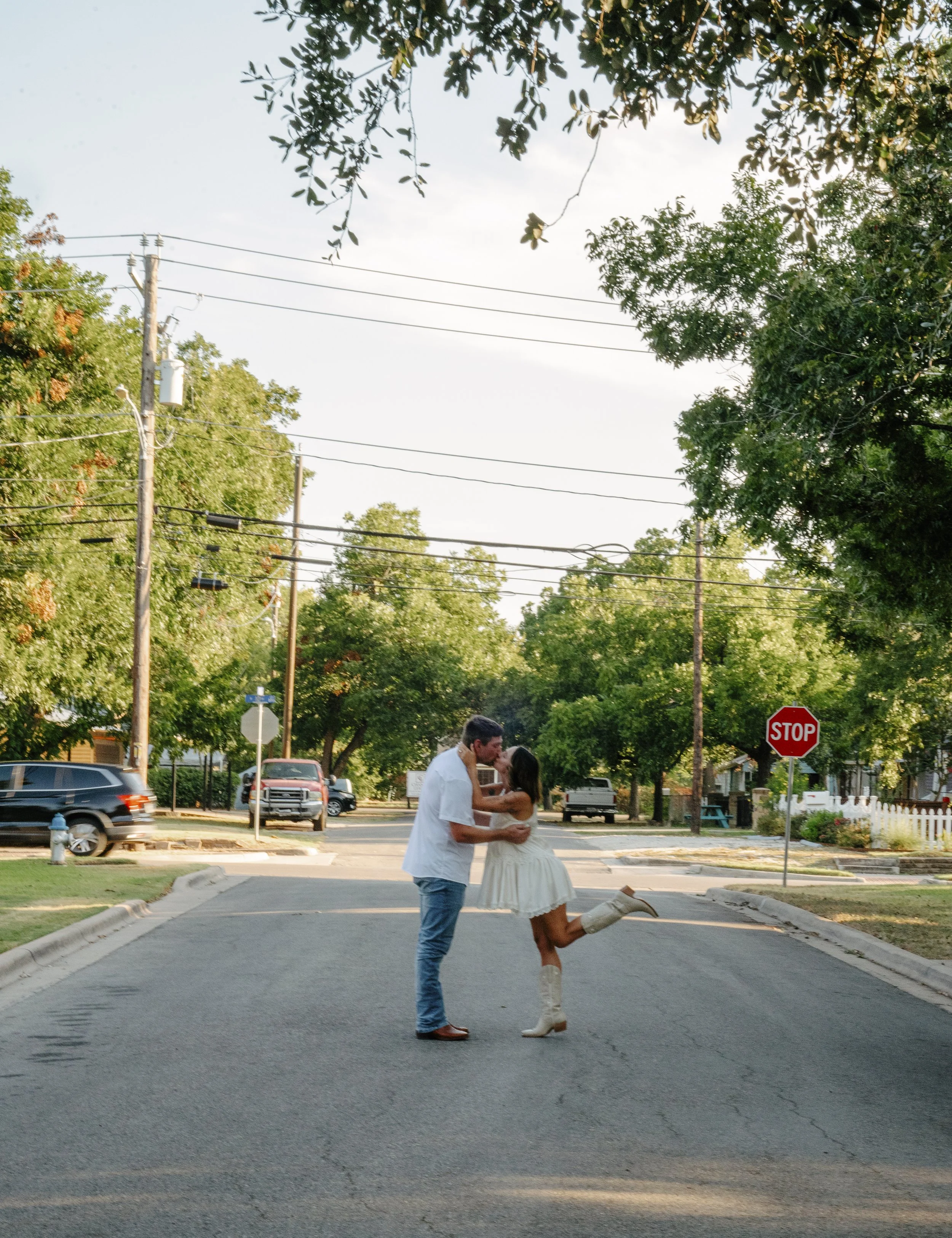 A couple kissing in the middle of the street during the day with trees and parked cars in the background.