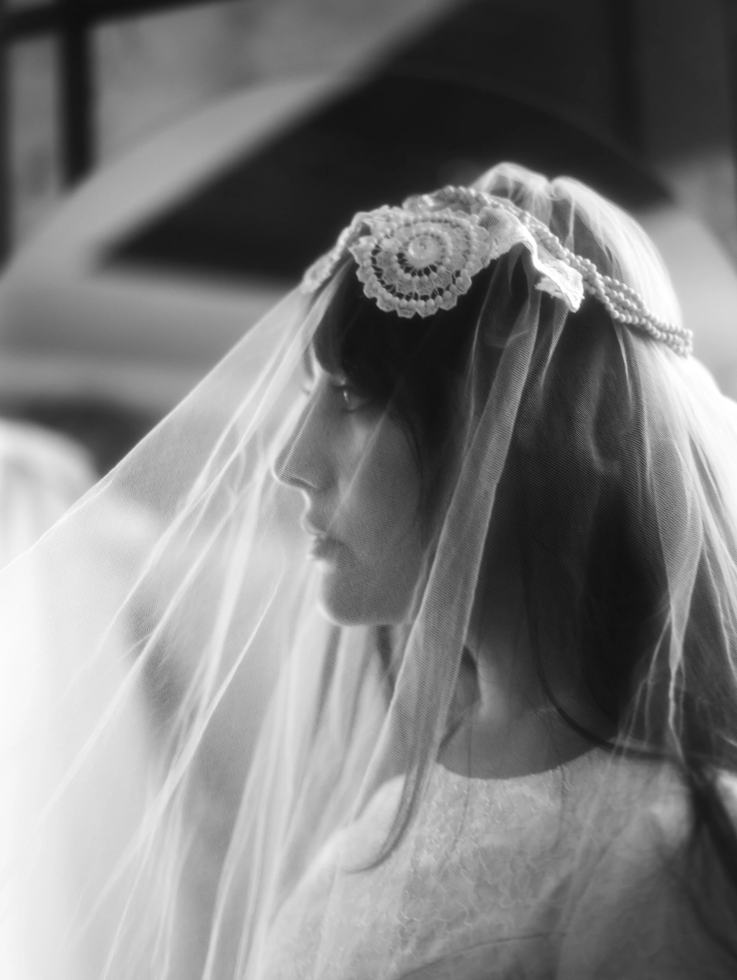Close-up black and white photo of a woman's head wearing a wedding veil with intricate lace detailing, blurred background.