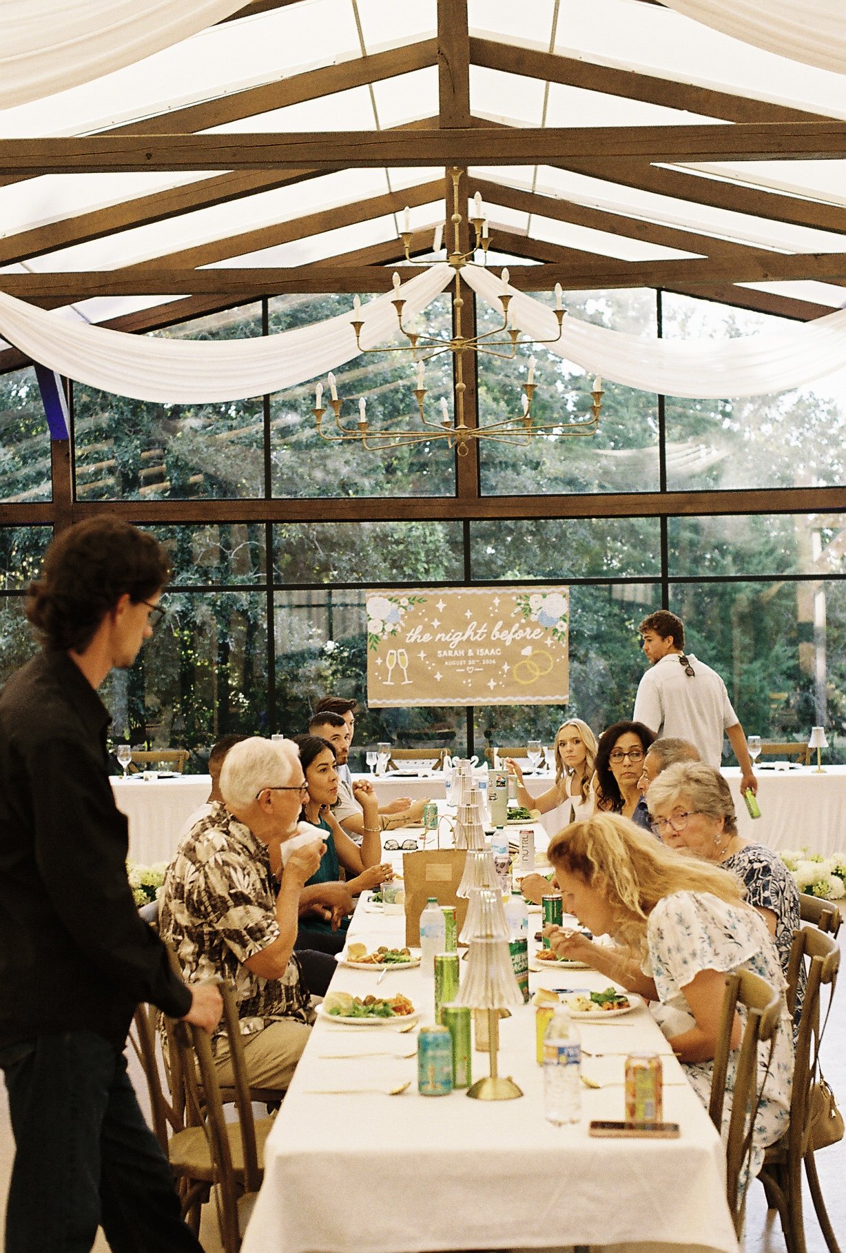 A wedding reception with guests seated at a long banquet table, servers attending to them, and a sign in the background reading 'the night before' with wedding details for Sarah and Isaac. Decor includes white draped fabric and a chandelier.