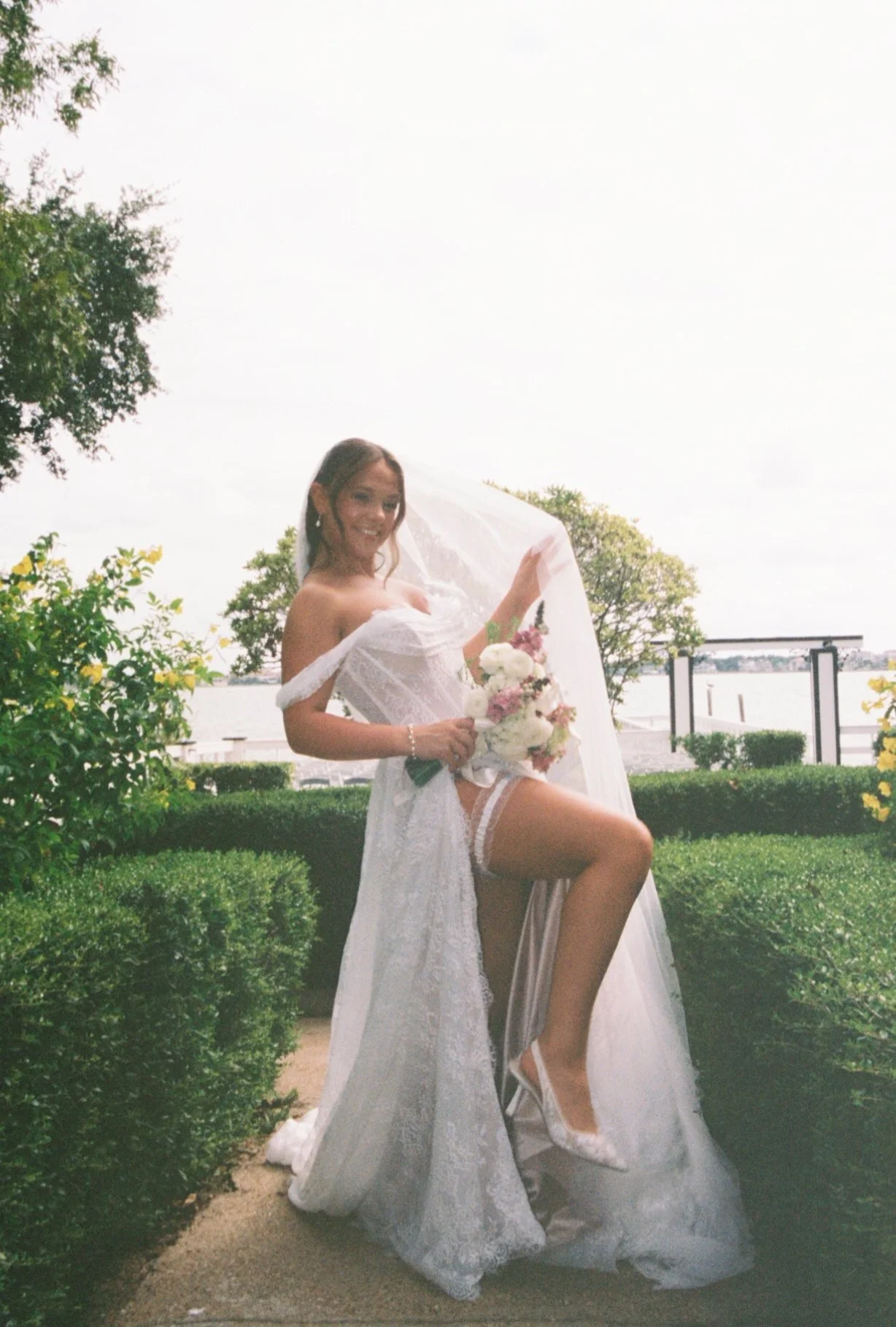 A smiling woman in a wedding dress holding a bouquet of flowers outdoors near a body of water with trees and a railing in the background.
