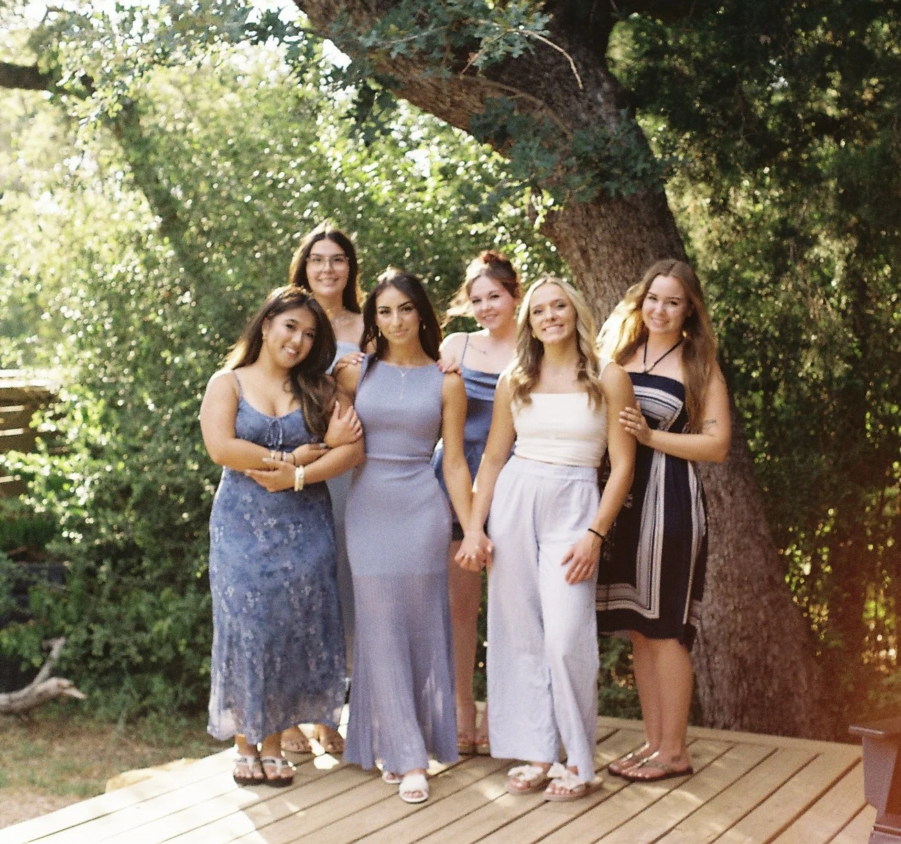 Six women standing outdoors on a wooden deck in front of a large tree, smiling and holding hands, dressed in summer dresses.