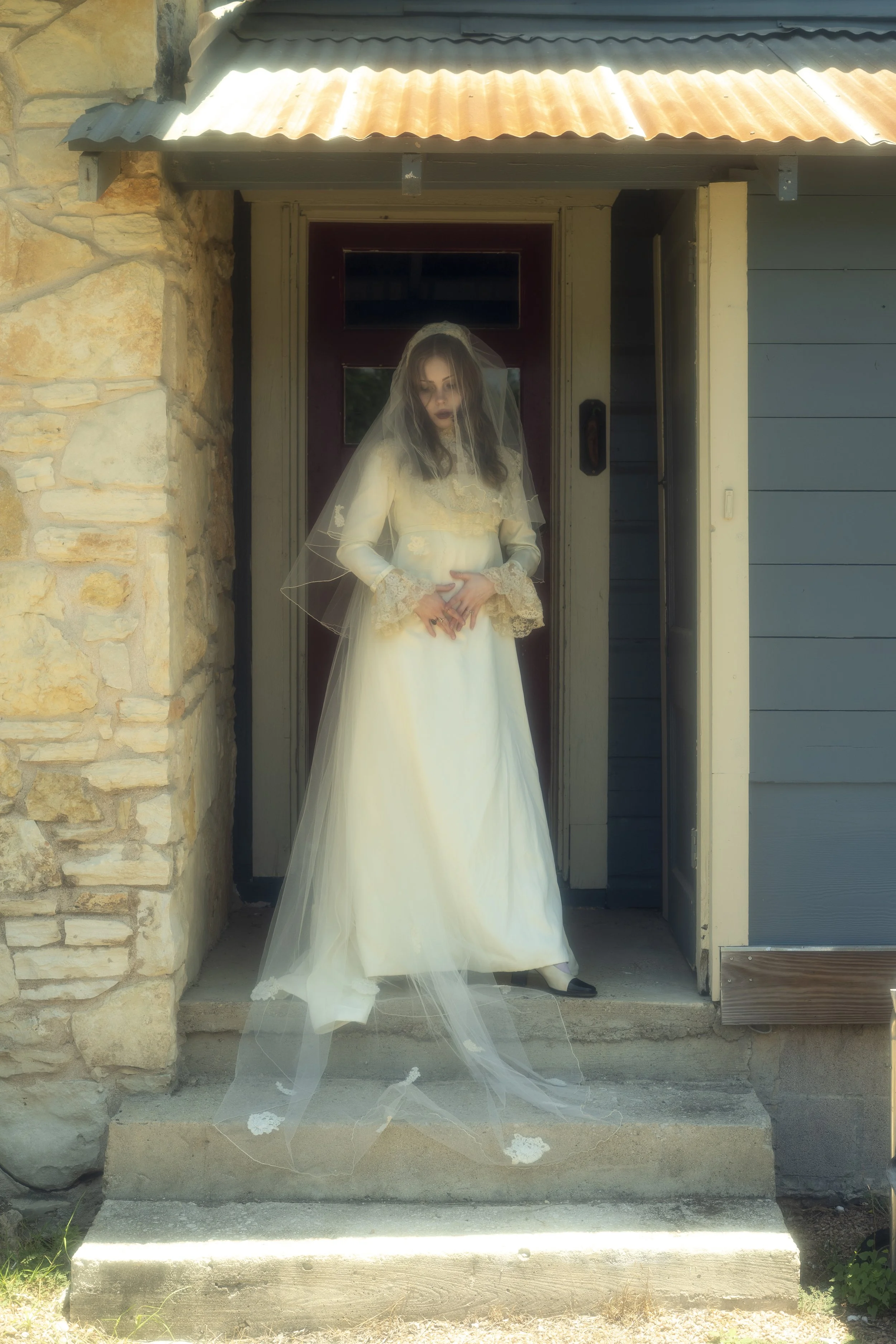 Woman in vintage wedding dress with lace sleeves and veil standing on front steps of stone and wood house.