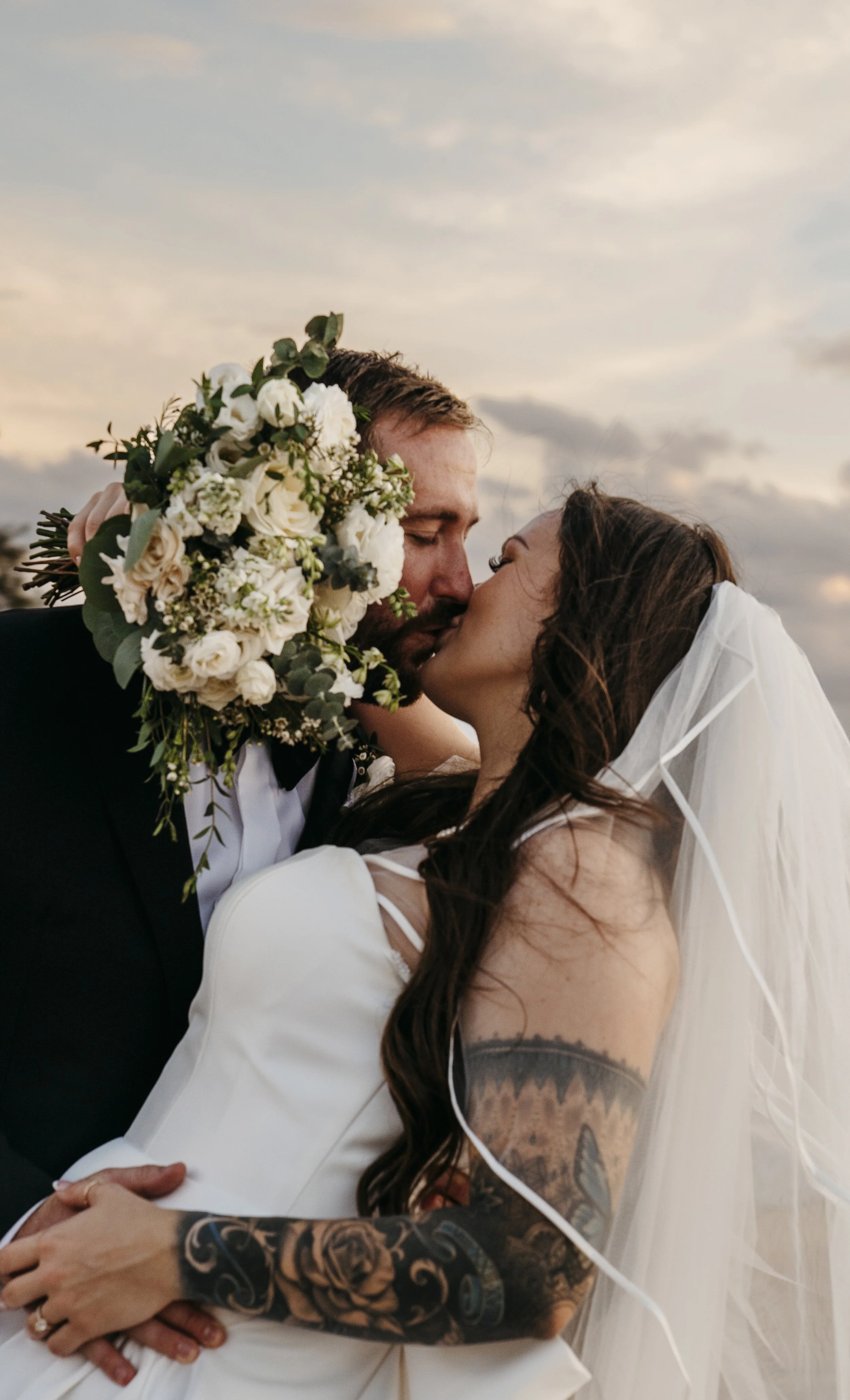 A couple sharing a kiss on their wedding day, with the bride wearing a veil and white dress and the groom holding a bouquet of white flowers, against a sunset sky background.