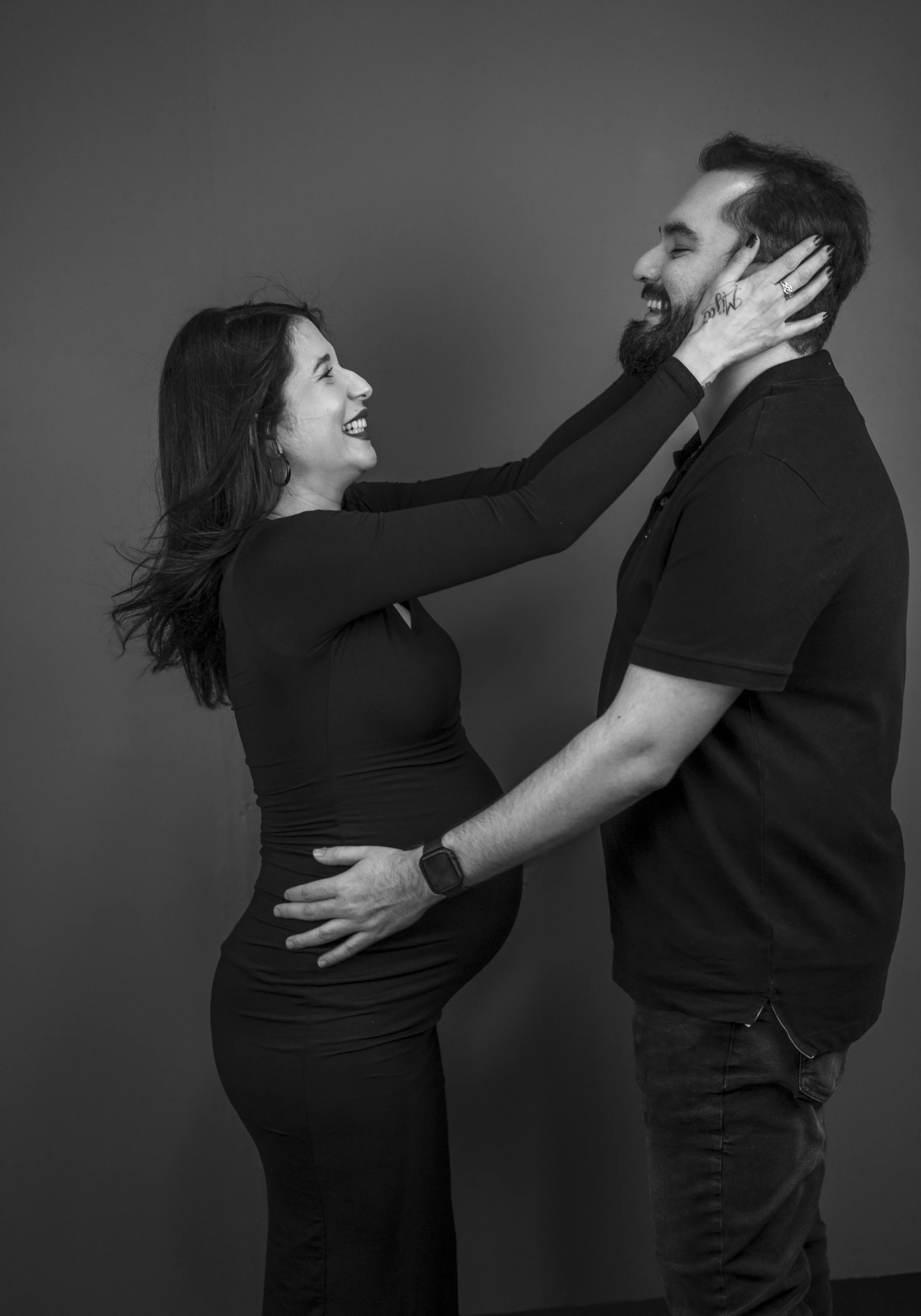 A pregnant woman and a man are smiling and looking at each other, with the woman touching the man’s face and the man holding her waist, against a plain gray background.