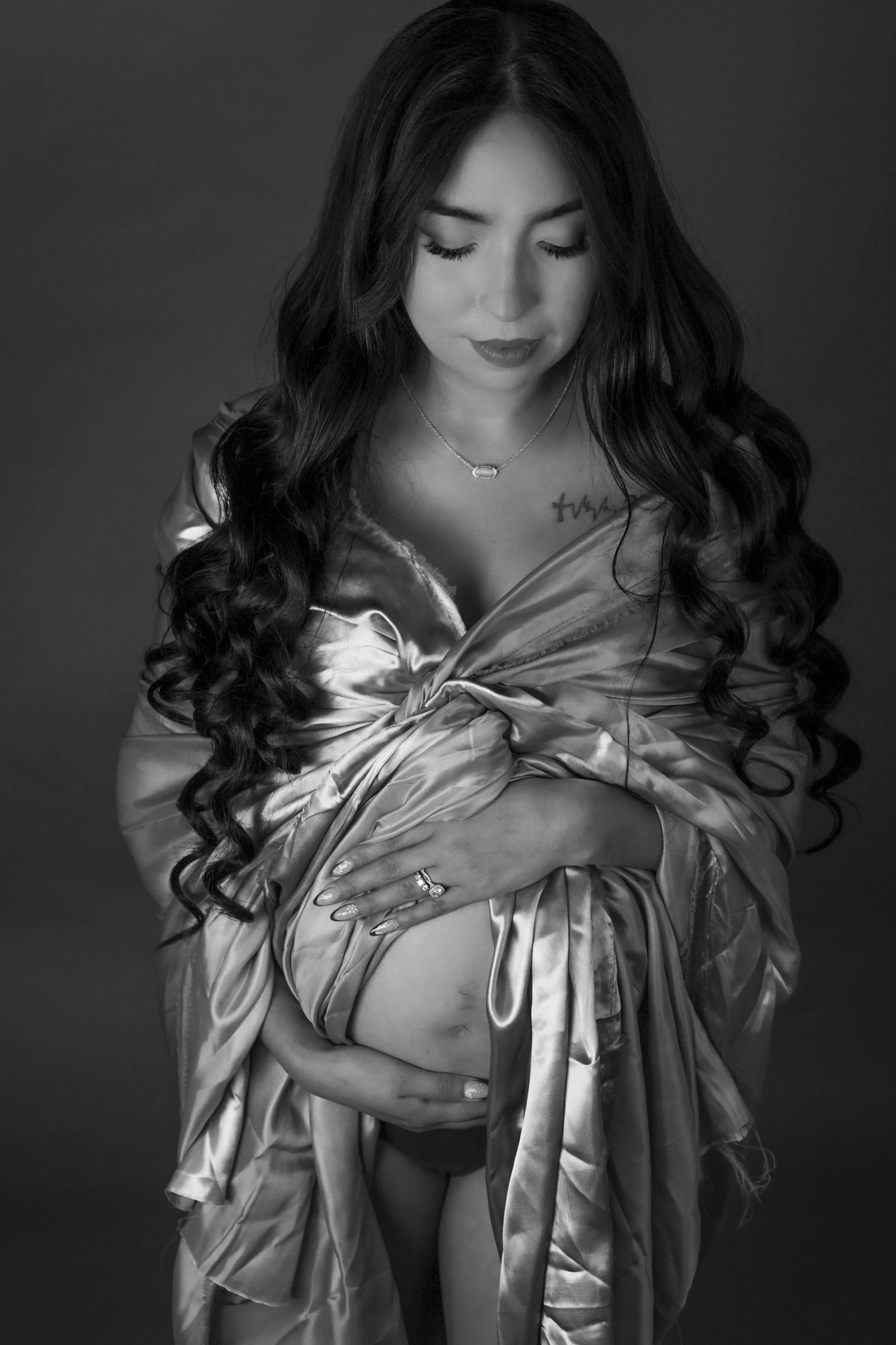 Black and white portrait of a woman with long, curly hair, wearing a satin robe, gently holding her pregnant belly with a serene expression and closed eyes.