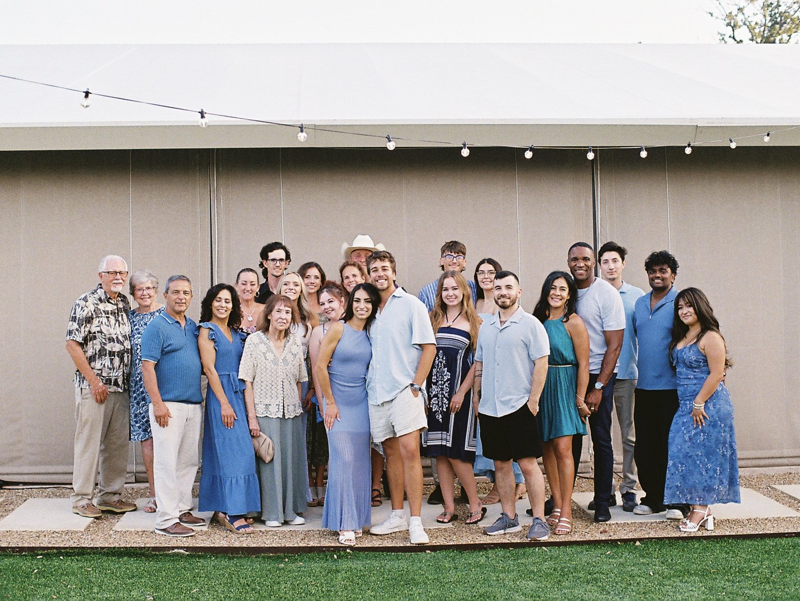 Group of people posing outdoors, standing in front of a beige wall and string lights, with grass in the foreground.