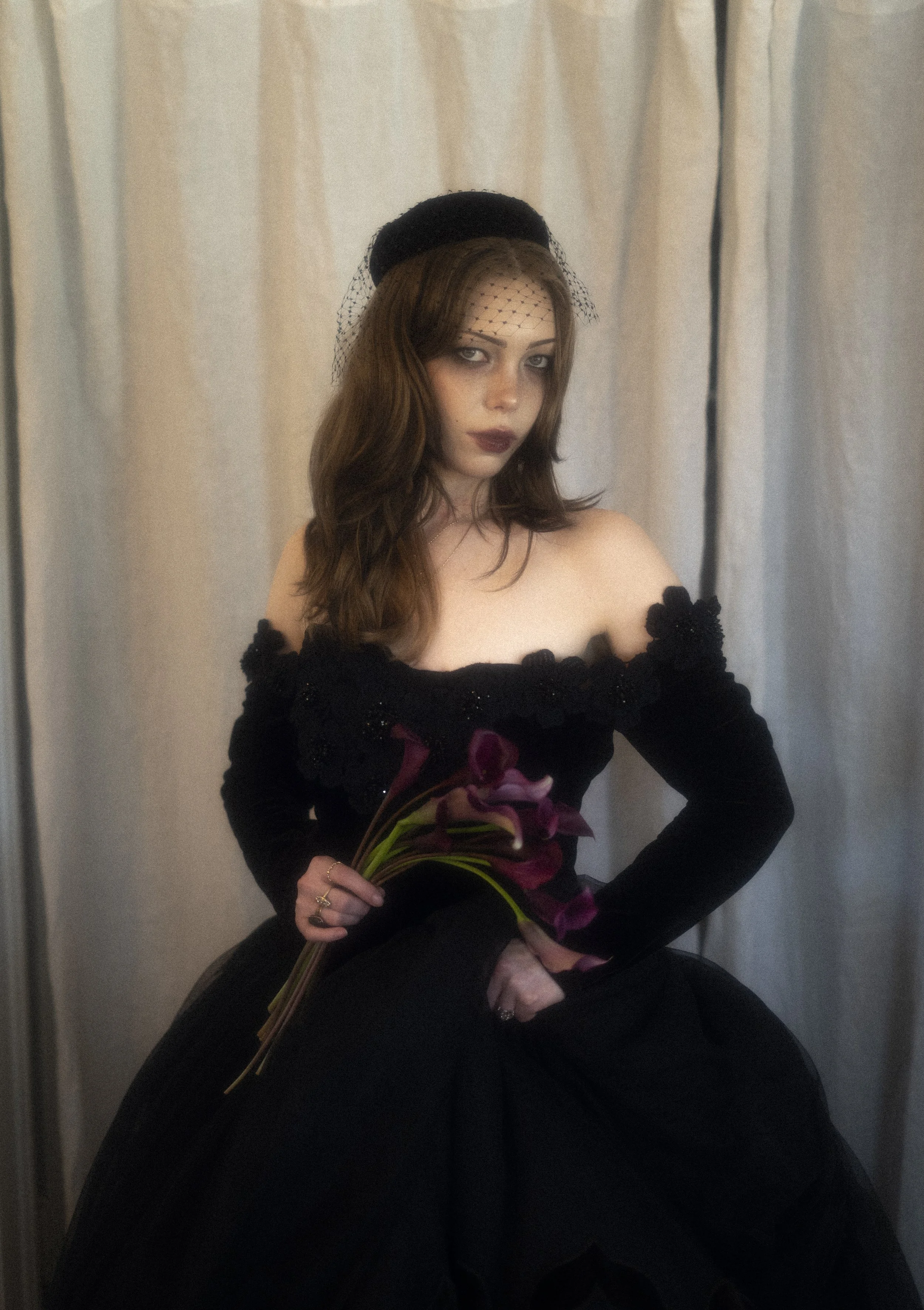A woman with long brown hair, wearing a black dress with lace details, black headband with netting, and dark makeup, holding a bouquet of dark pink and purple flowers, sitting in front of a beige curtain.
