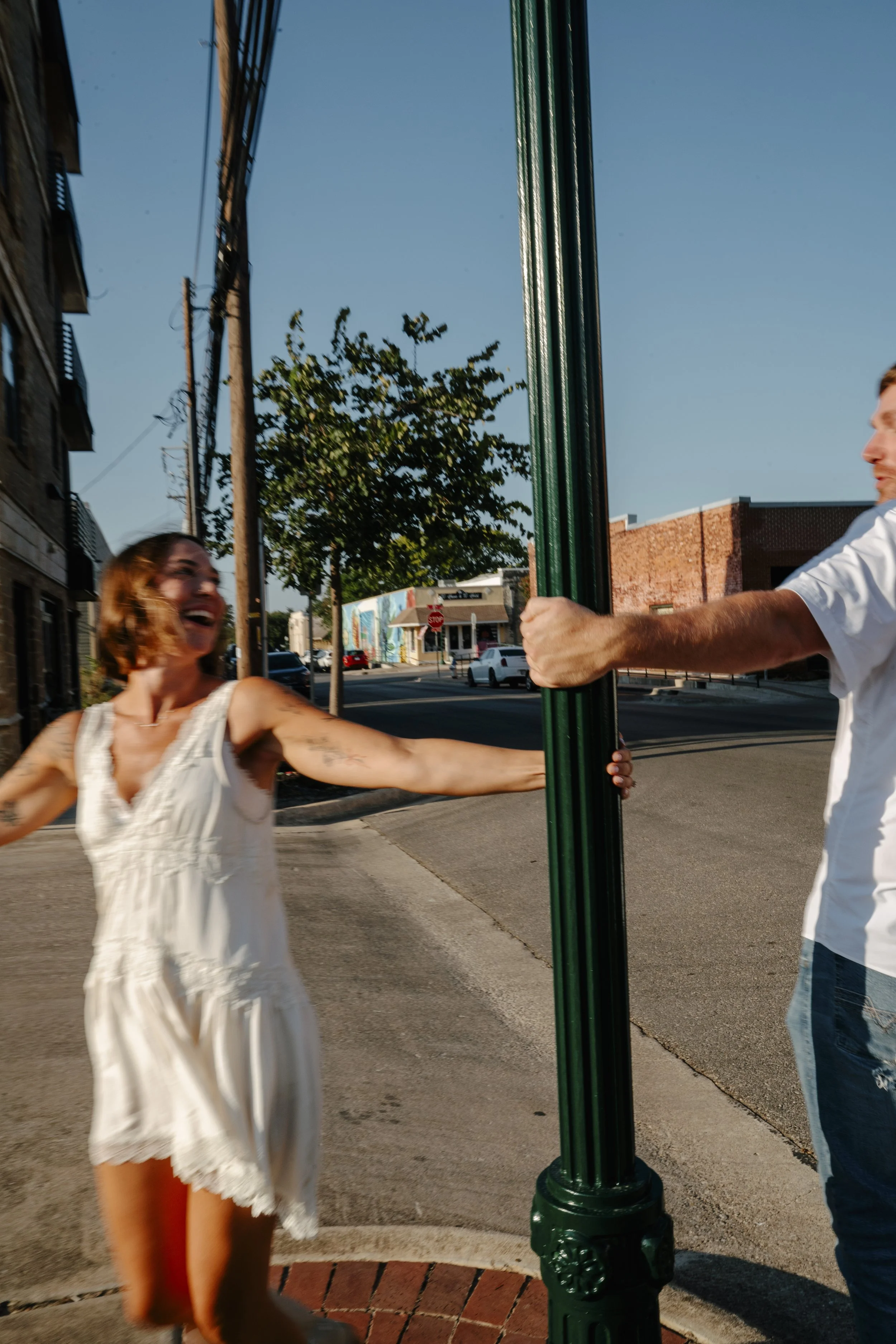 A woman in a white summer dress is dancing and smiling, reaching out her arm, while a man in a white shirt holds a green pole on a city sidewalk under a clear blue sky. Trees and buildings are visible in the background.