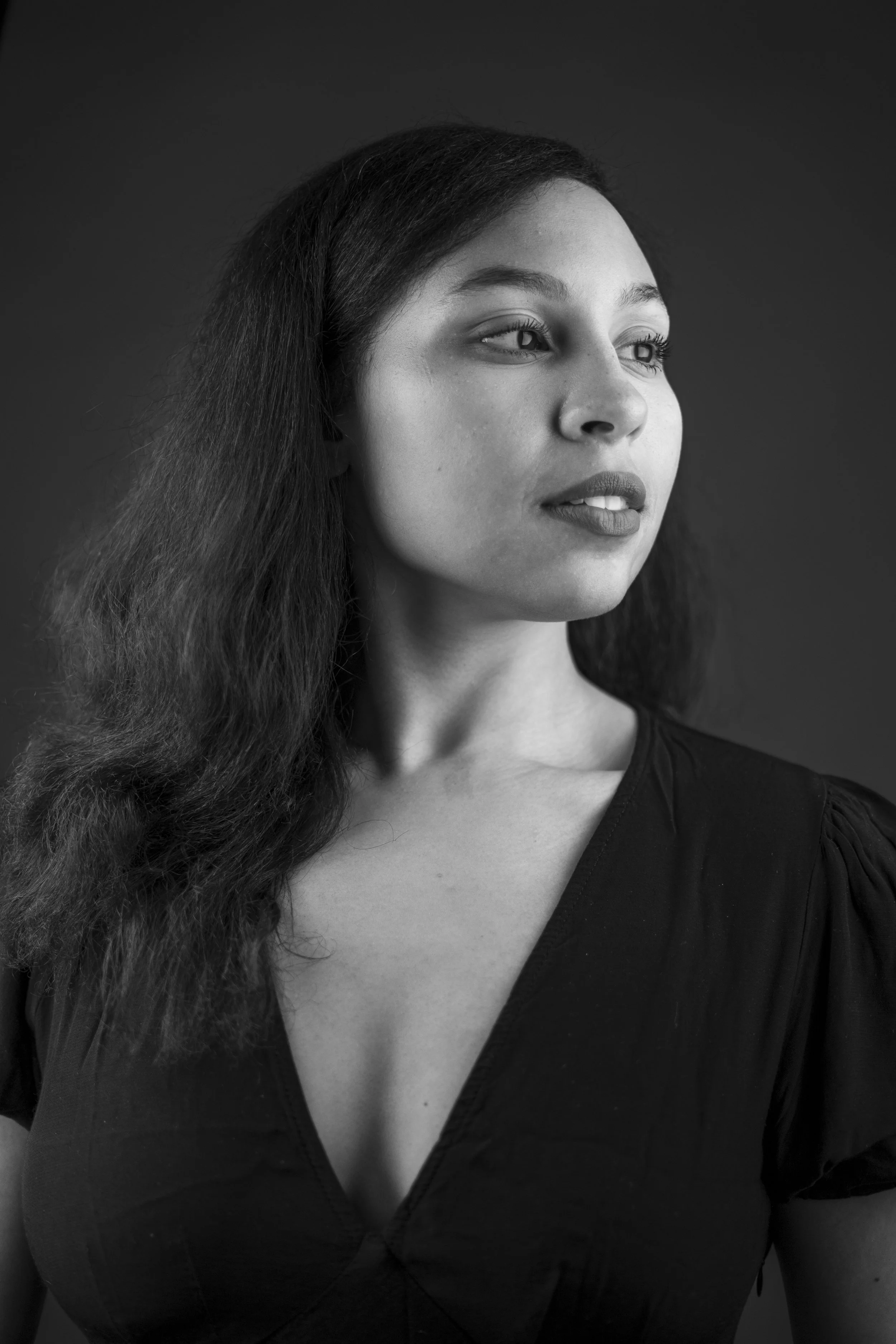 A black-and-white portrait of a woman with long, wavy hair, looking slightly to her right, wearing a dark V-neck top.