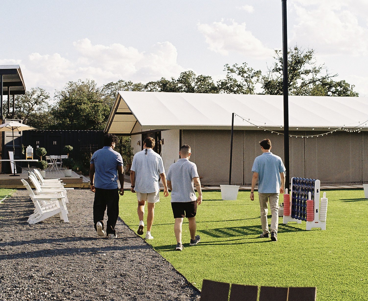 Four young men walking on a gravel path toward a gray building with string lights, surrounded by outdoor furniture, a grassy area, and decorative items, in a backyard setting under a partly cloudy sky.