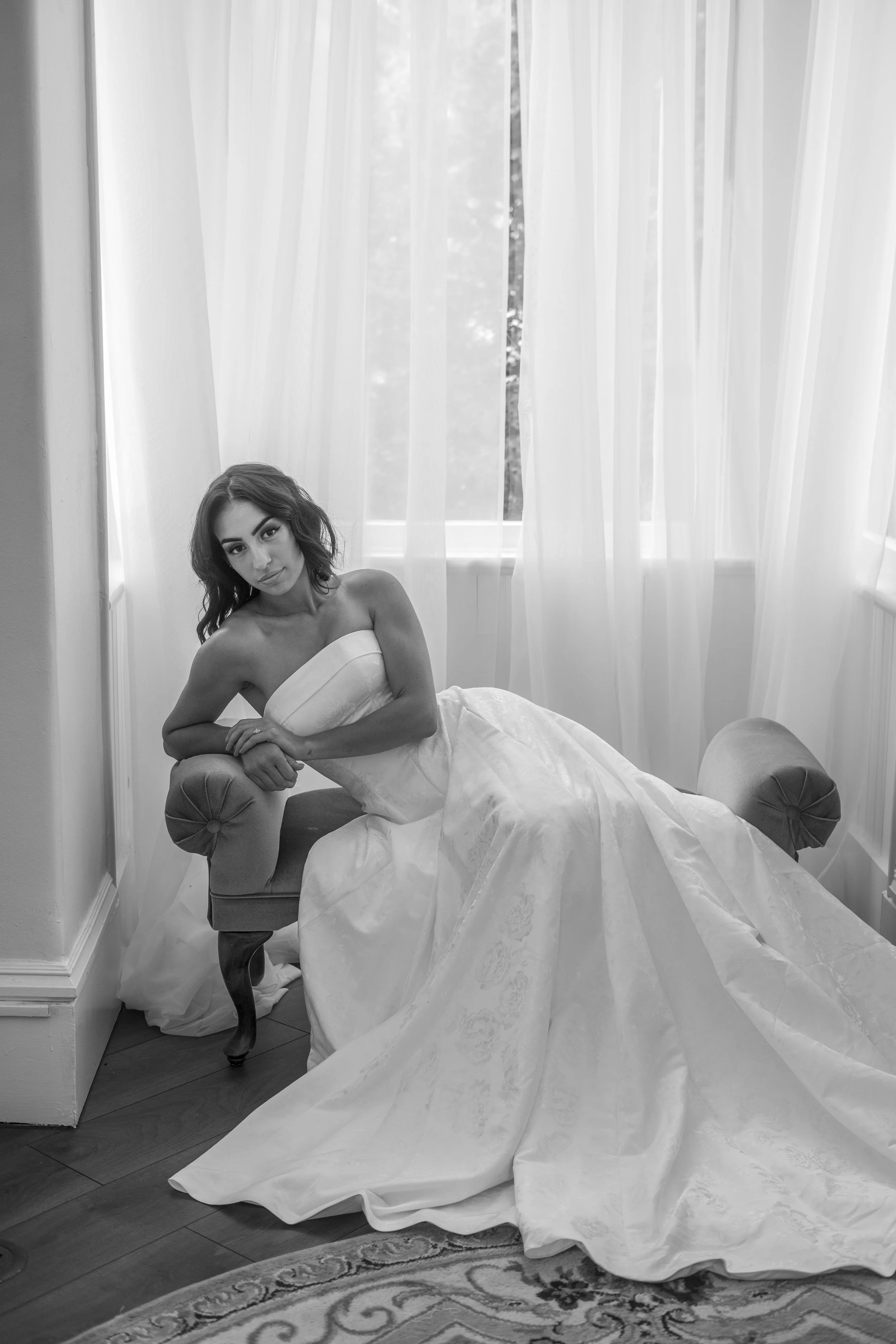 A woman in a wedding dress sitting on an ottoman in a room with large windows and white curtains.