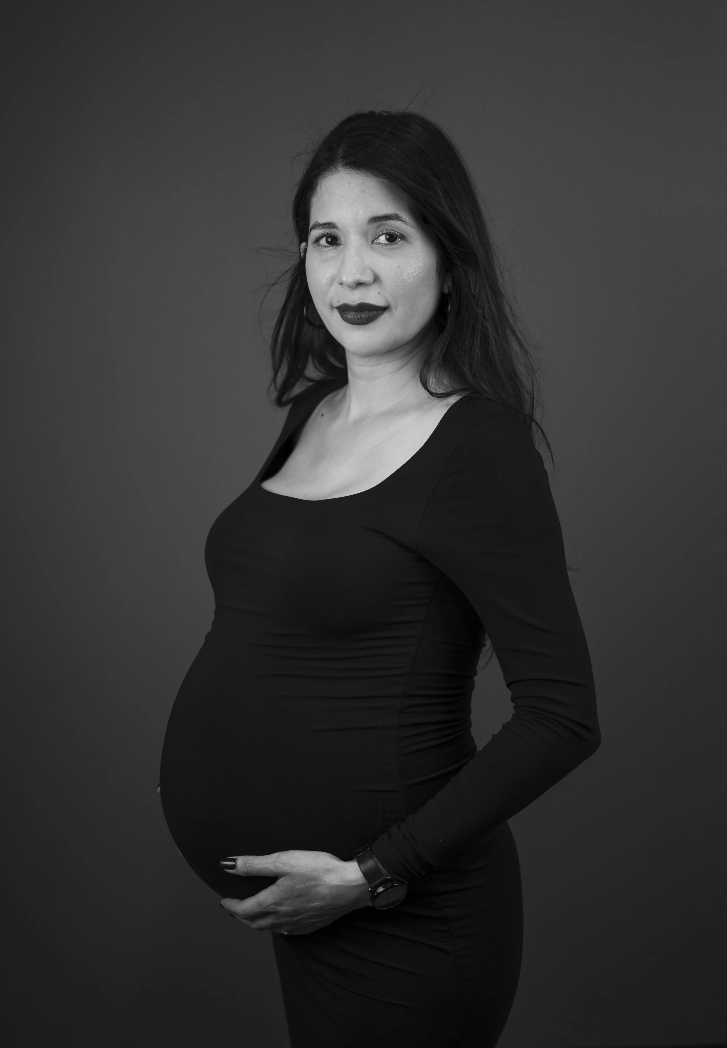 Black and white portrait of a pregnant woman with dark, shoulder-length hair, wearing a fitted black long-sleeve dress, standing in front of a plain background.