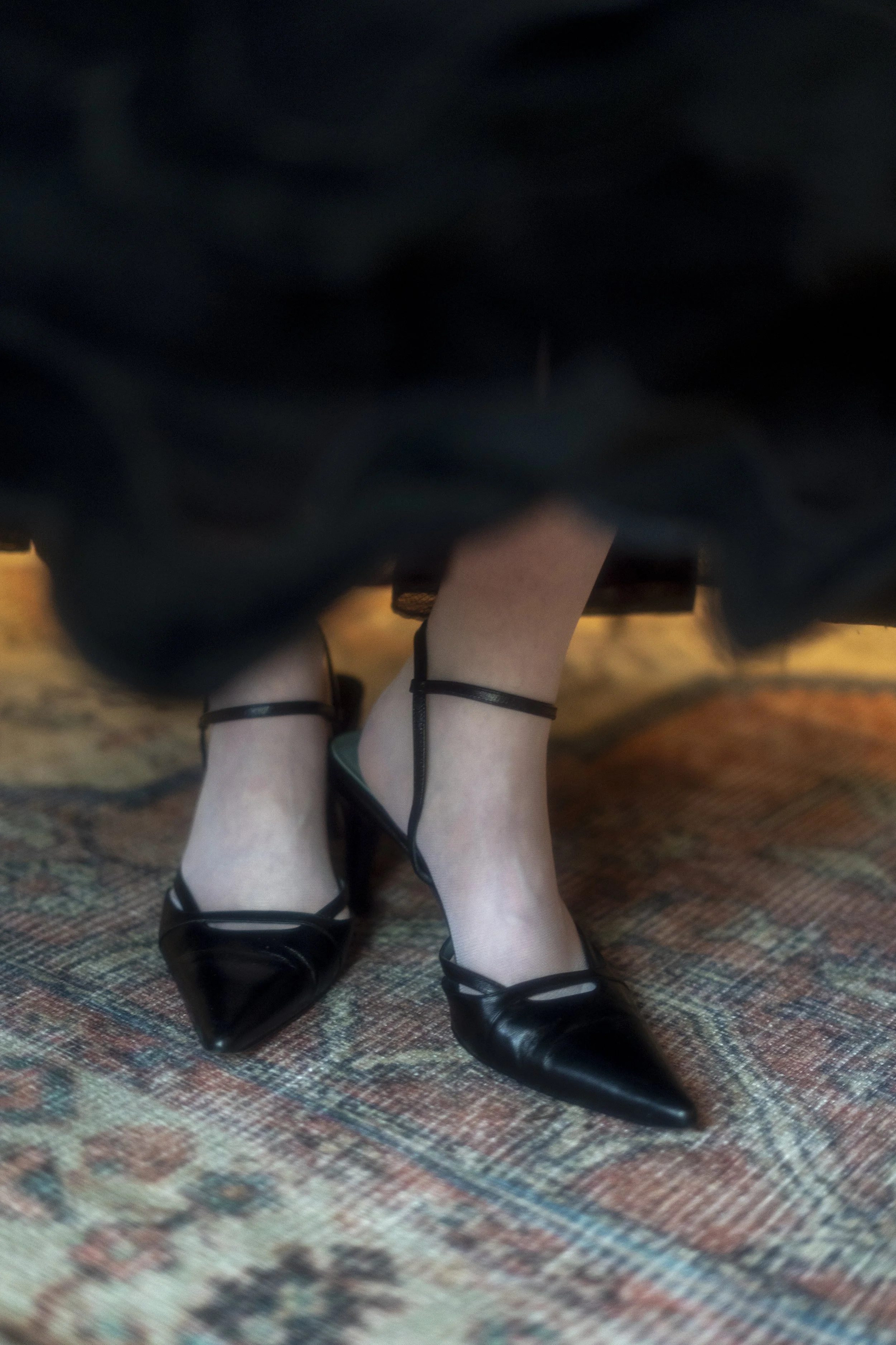 Person wearing black heeled shoes and sheer black stockings, sitting on a patterned carpet with visible foot and ankle accessories.