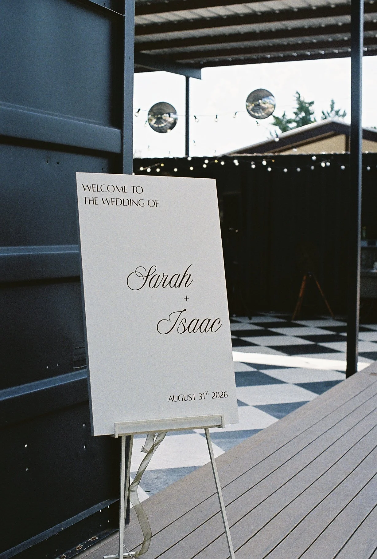 Wedding welcome sign with names Sarah and Isaac, dated August 31, 2026, on an easel at an outdoor venue with string lights in the background.