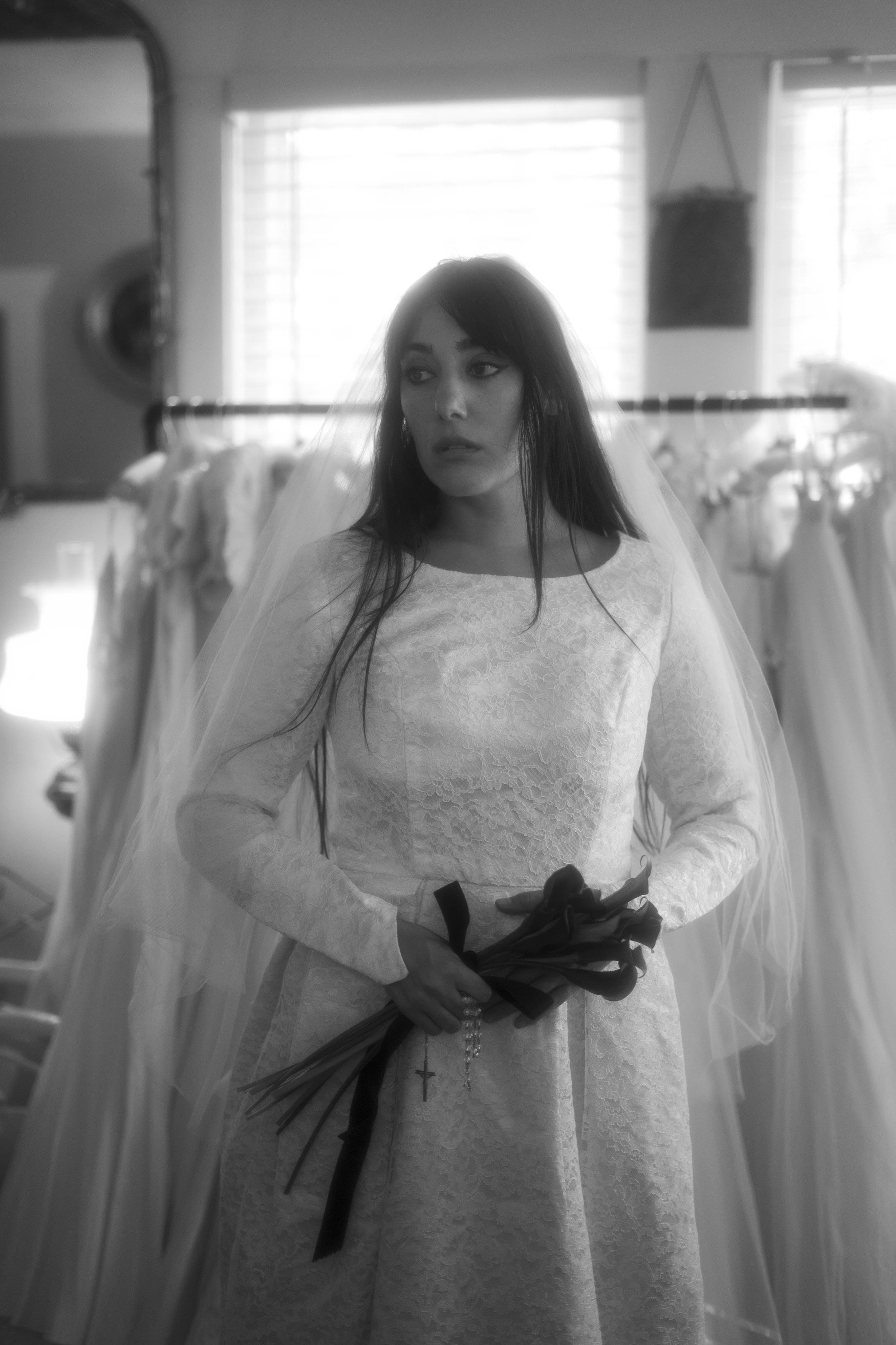 A woman dressed in a wedding gown holding a bouquet of flowers, standing inside a bridal boutique with wedding dresses hanging in the background.