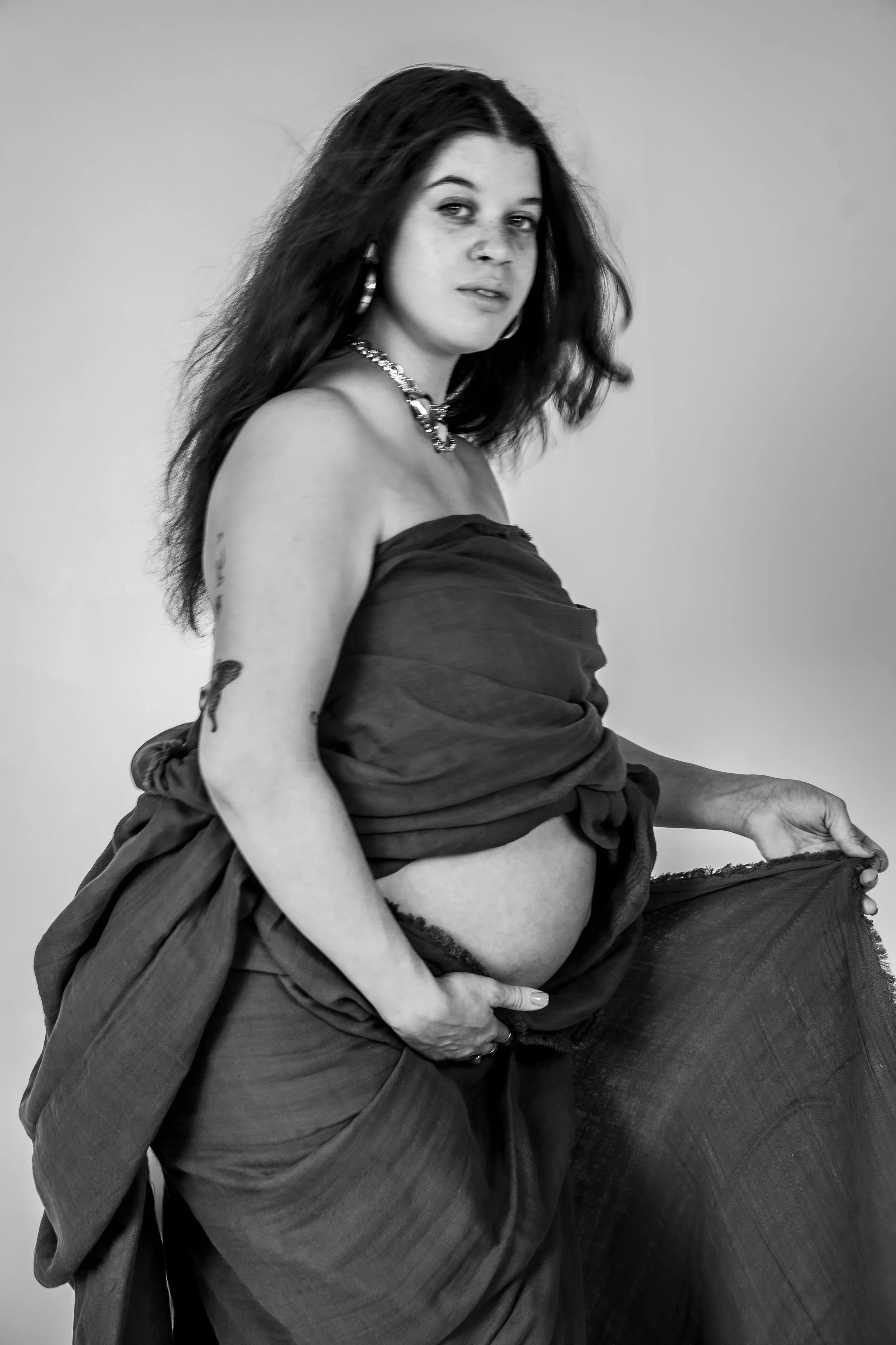Black and white photo of a woman with dark, wavy hair wearing jewelry and a loose, draped fabric around her shoulders and waist, partially lifted to reveal her midriff.