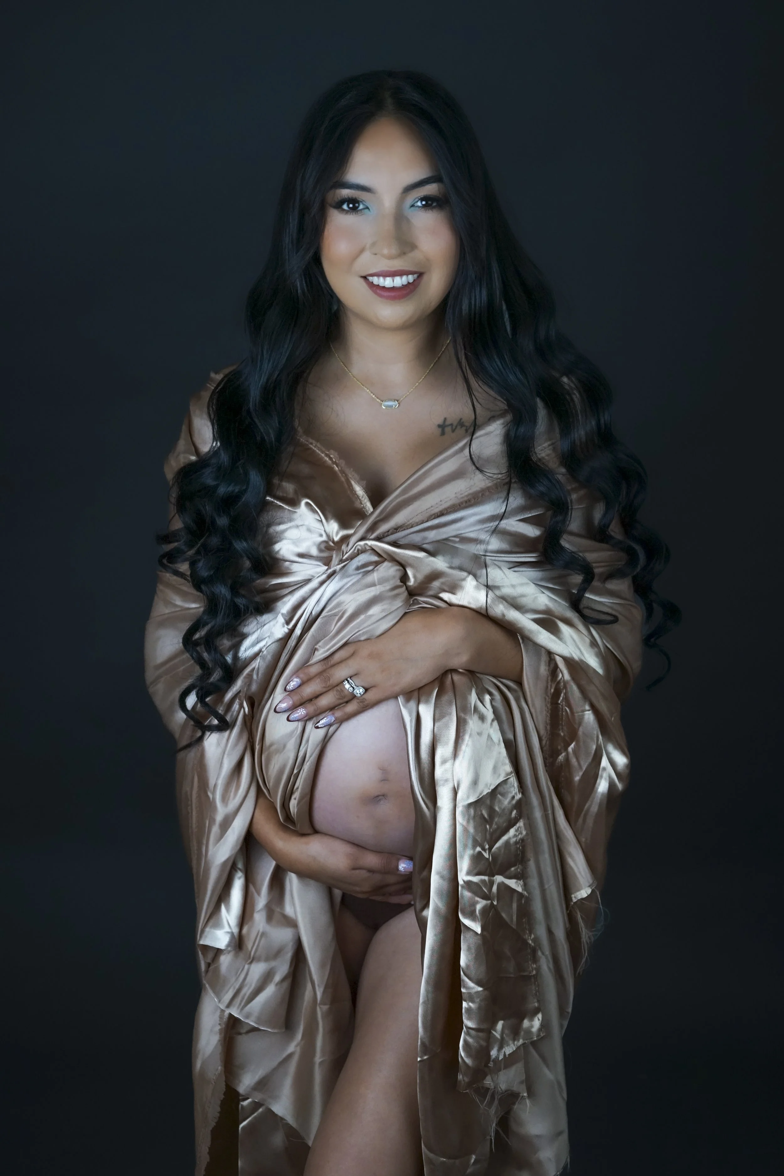 A pregnant woman with long black curly hair, smiling, wearing a satin beige robe, standing against a dark background, holding her belly.