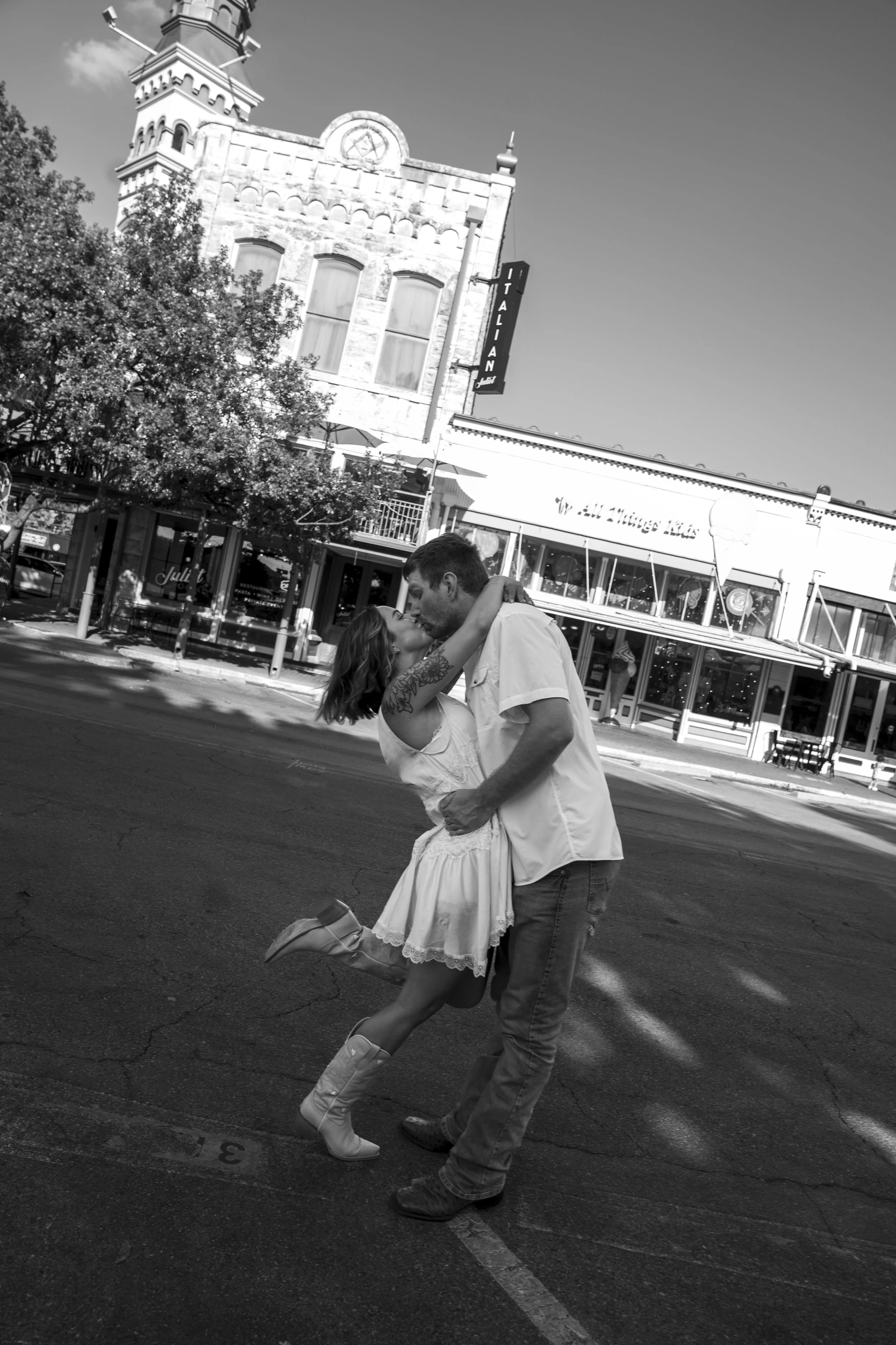 A couple dance passionately on a city street at night. The woman, wearing a knee-length dress and cowboy boots, lifts one leg as the man holds her. Behind them, there are trees, a building with a sign that reads 'ITALIAN,' and a restaurant with a str