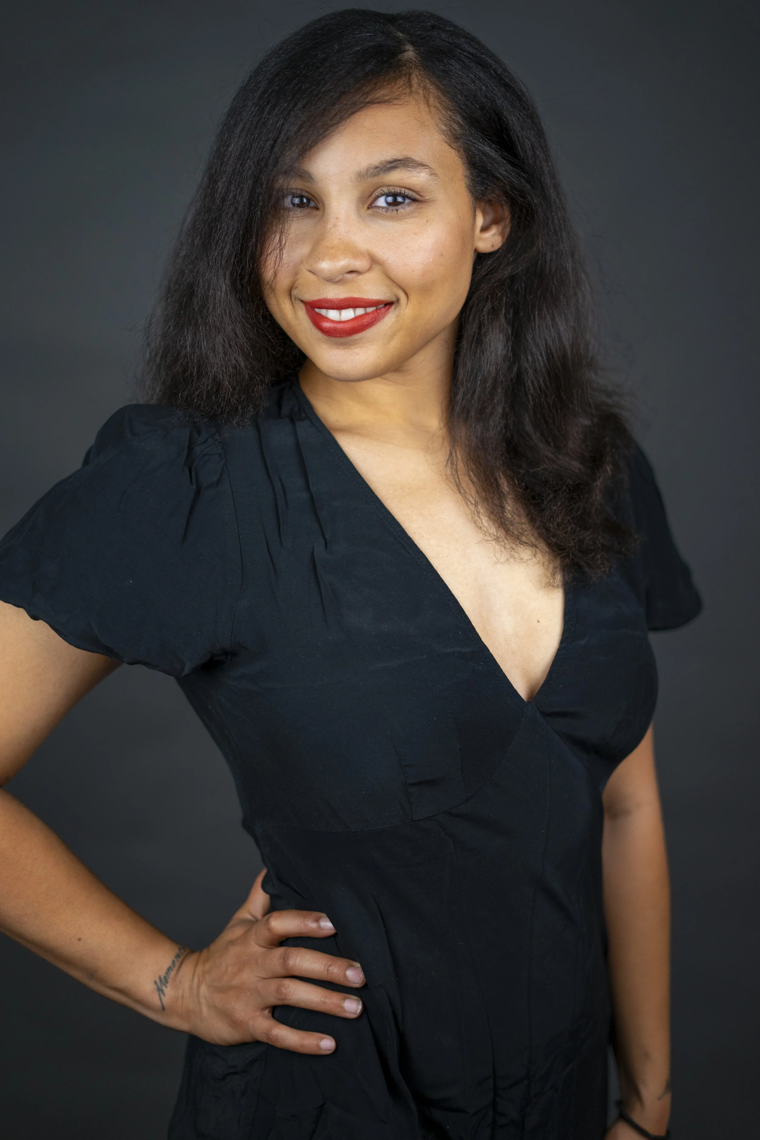 A woman with medium skin tone, black wavy hair, and red lipstick, smiling, wearing a black short-sleeved dress, standing against a dark gray background, with her left hand on her hip.