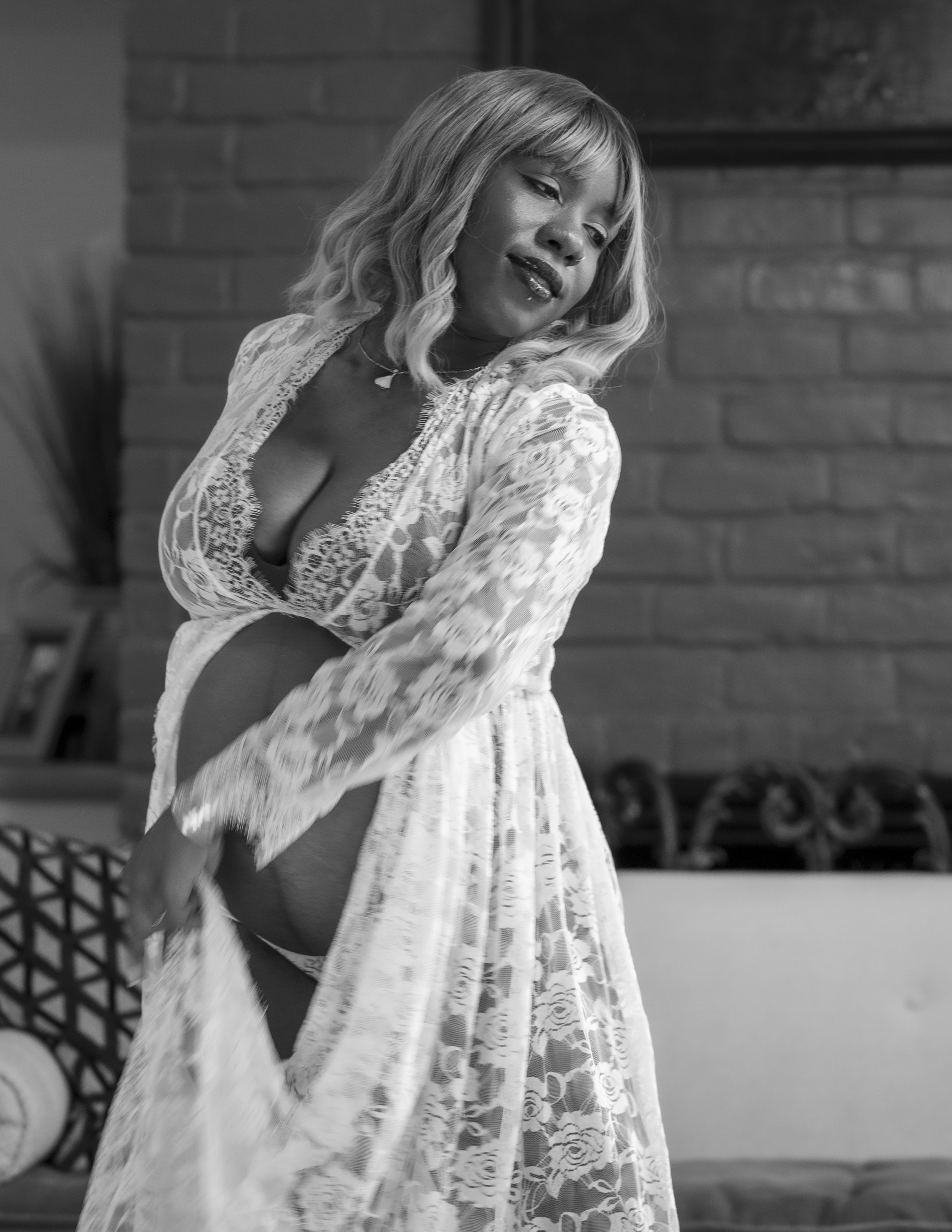 A woman with blonde hair in lace lingerie and robe, posing indoors against a brick wall.