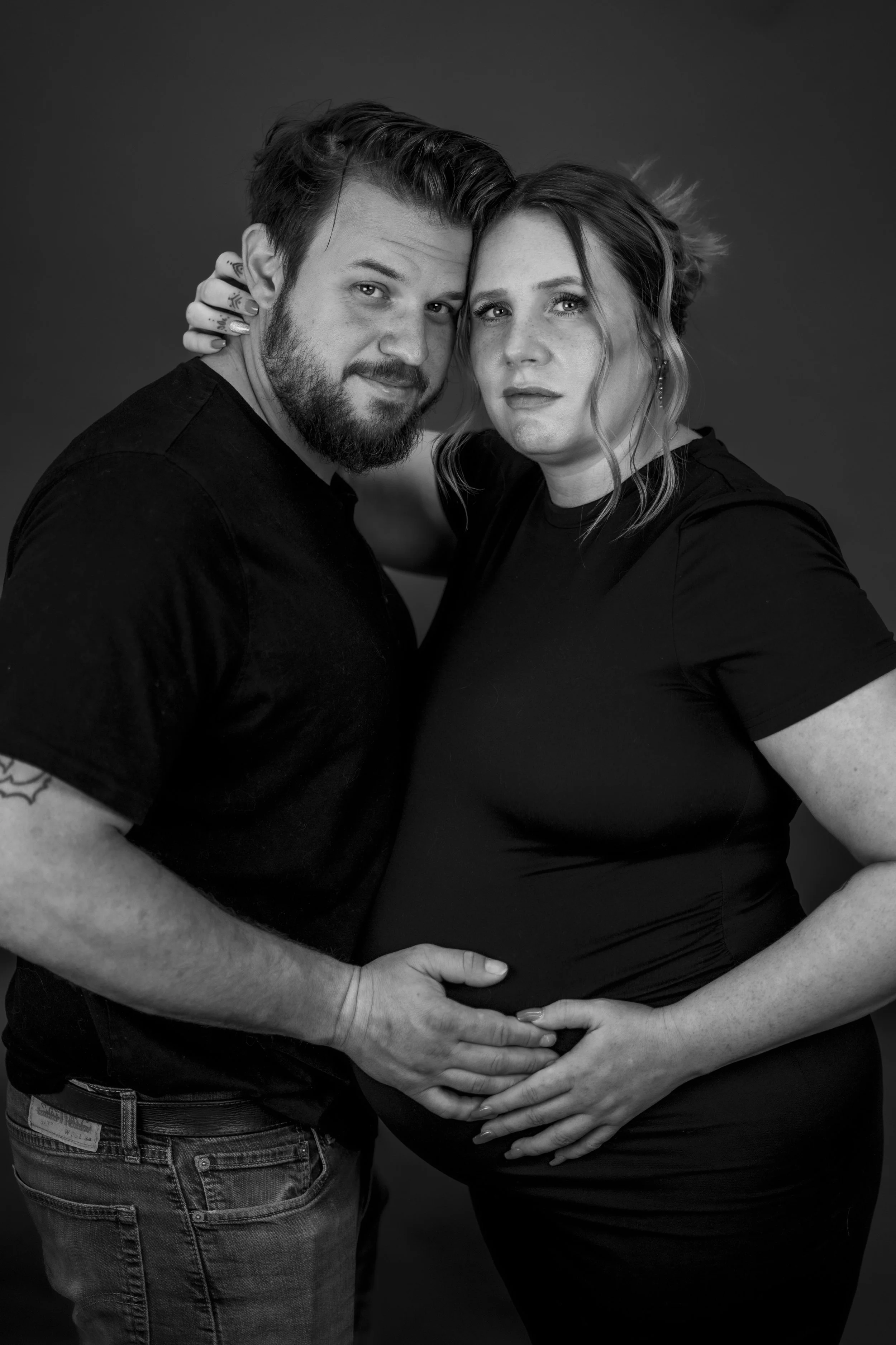 A couple, with the man having a beard and tattoos, and the woman with curly hair and earrings, posing closely together, with the man’s hand on her pregnant belly, in black and white studio portrait.