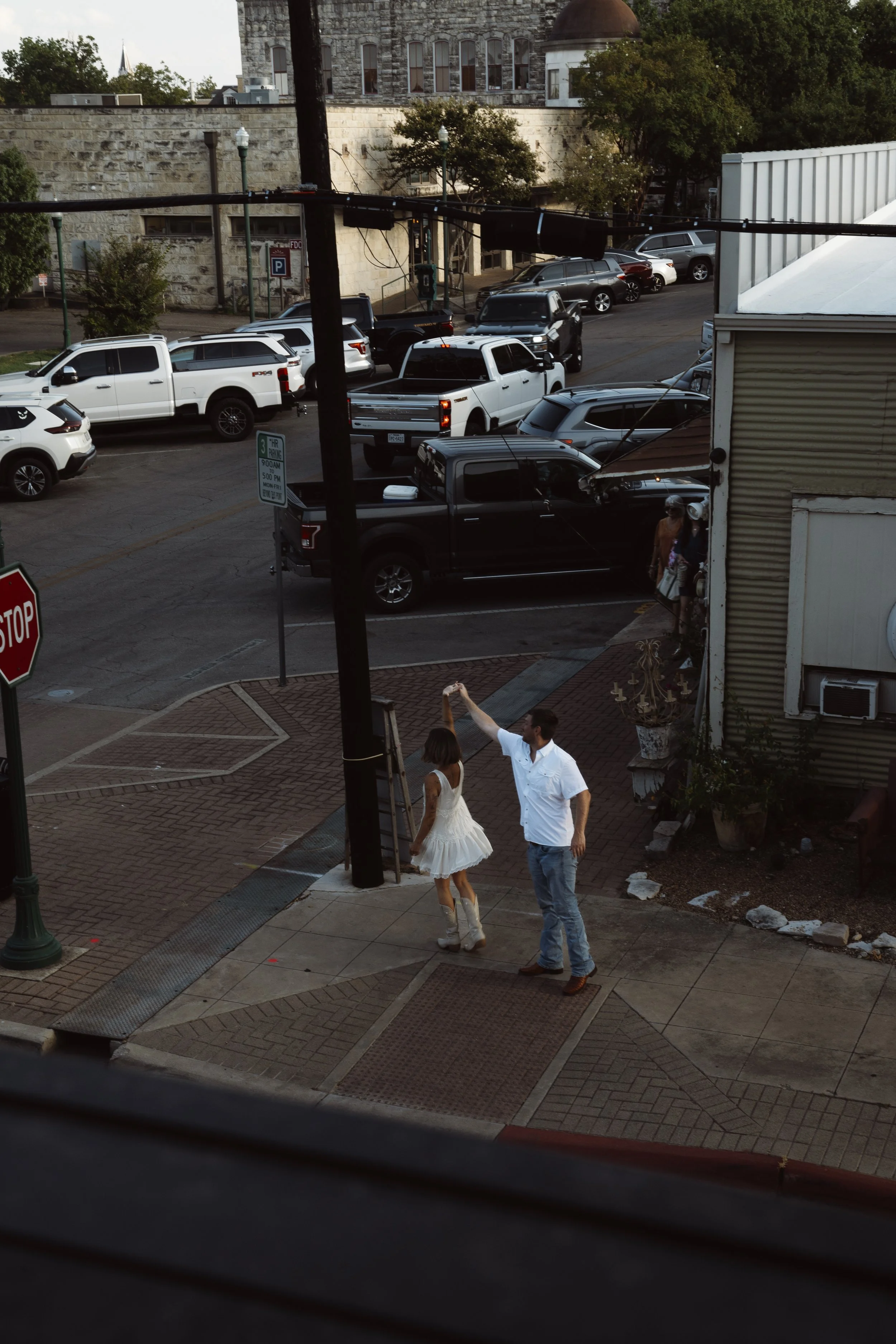 A street scene showing a couple walking on the sidewalk, with the woman in a white dress and the man in a white shirt and jeans. In the background, there are several parked cars and a building with a stone facade. There are street signs, lampposts, a