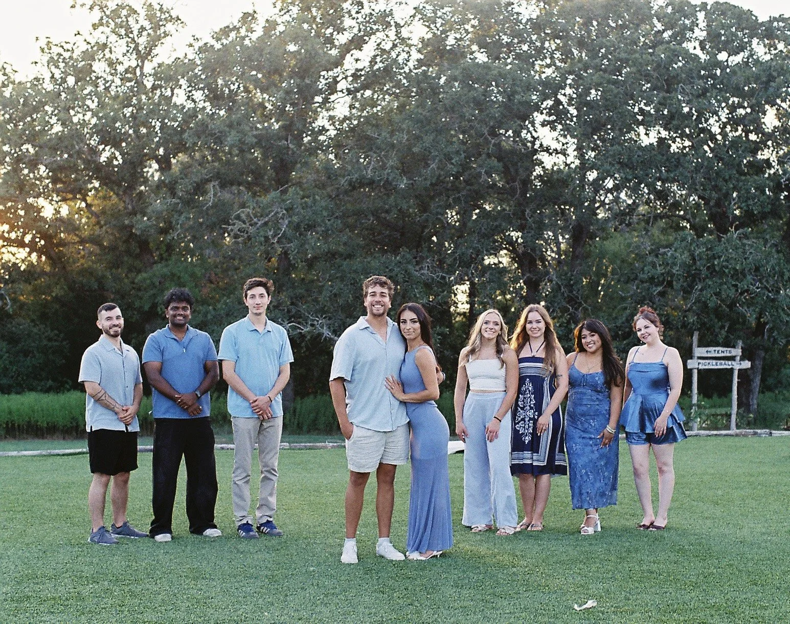 Group of nine young adults standing on a grassy field, smiling, with trees in the background and a sign that says 'Tents' and 'Fickleballe' in the distance.