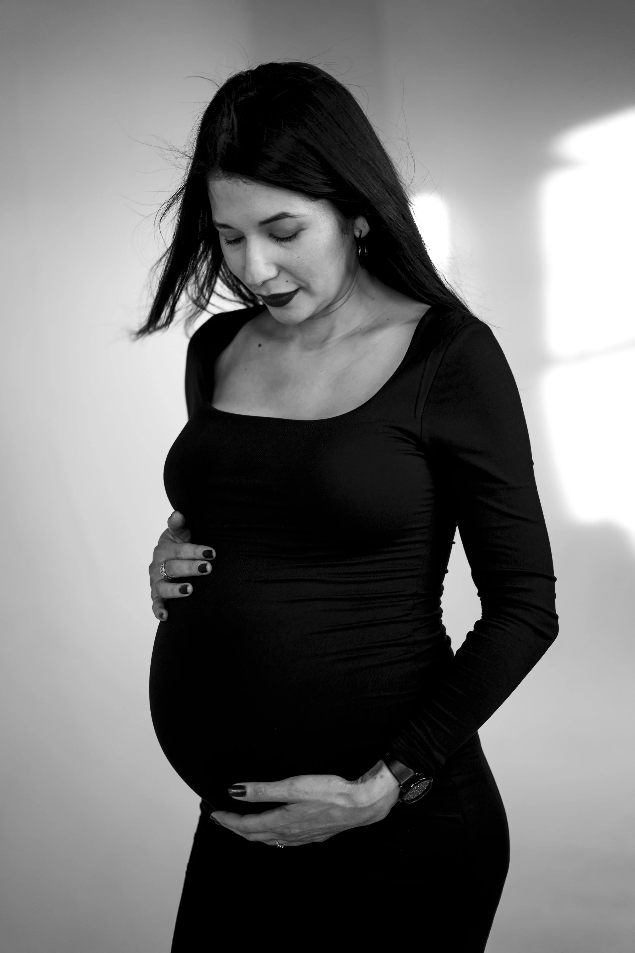 Black and white photo of a pregnant woman with dark hair, wearing a fitted black dress, cradling her baby bump and looking down.