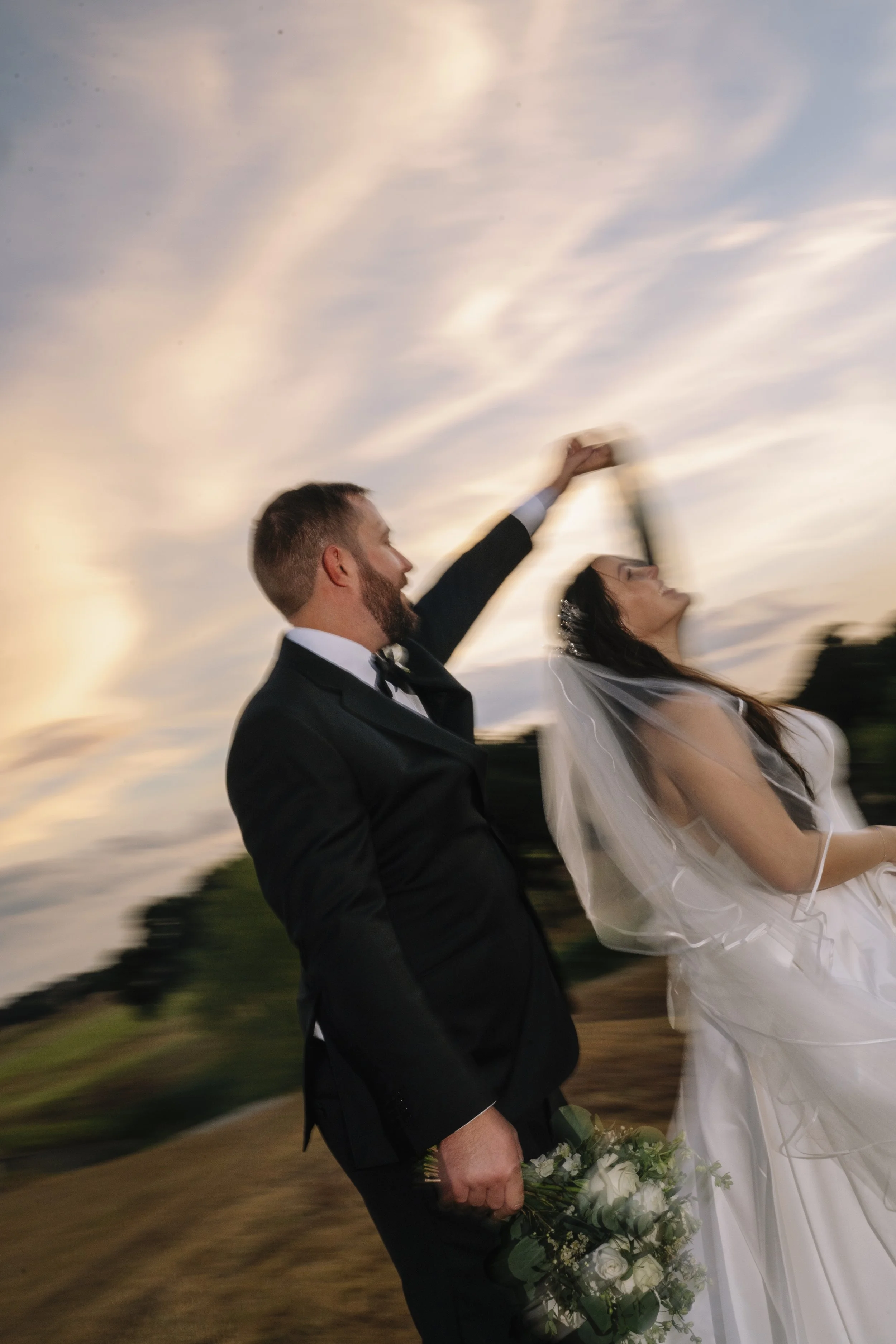 A newlywed couple dancing outdoors during sunset, with the groom in a black tuxedo and the bride in a white wedding gown, holding a bouquet of white flowers.